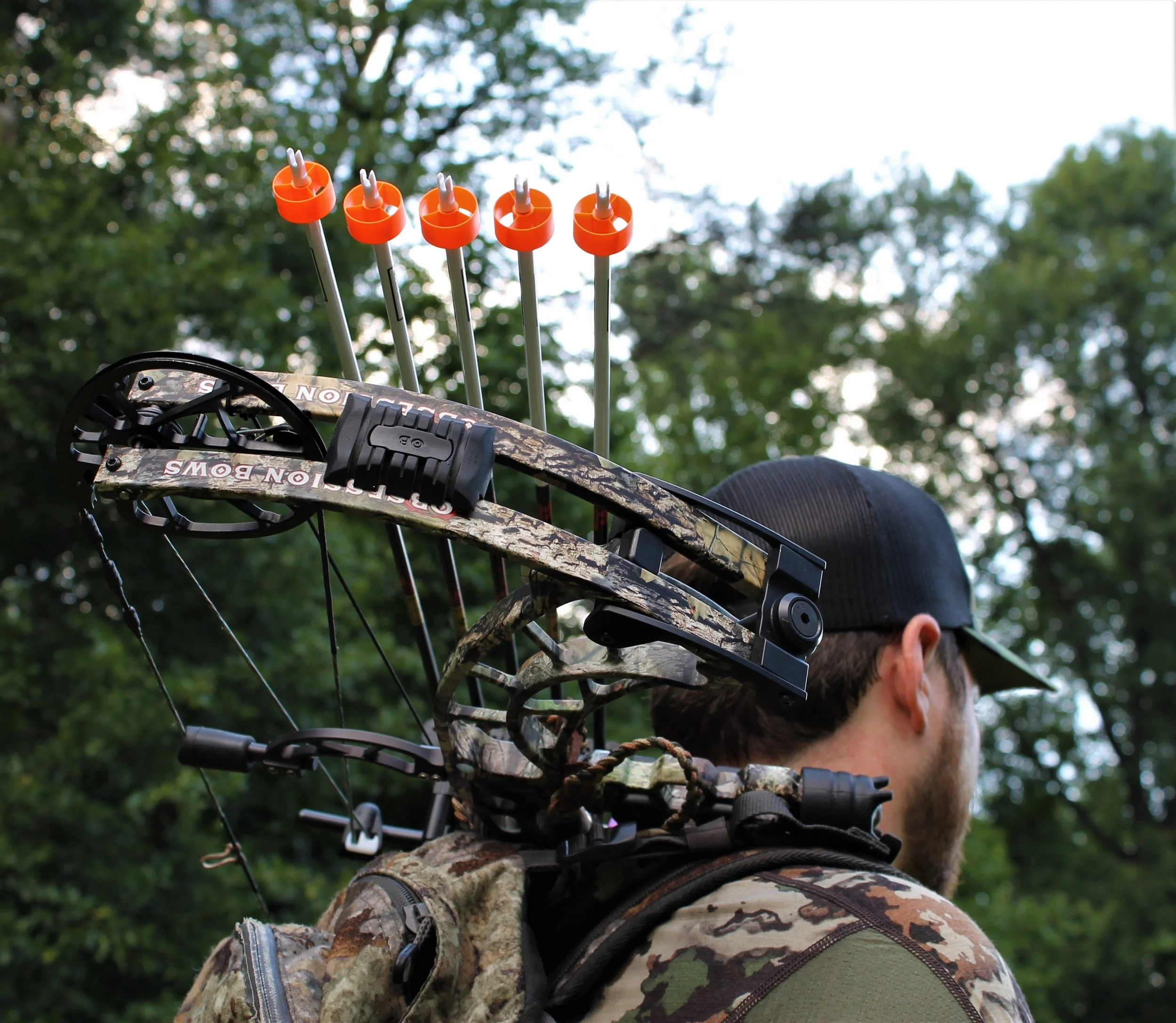 Close-up of a compound bow with five orange and white arrow nocks loaded, mounted on an outdoor hunter's camouflage camouflage, with a person wearing camouflage clothes and a cap in the background amidst green trees.
