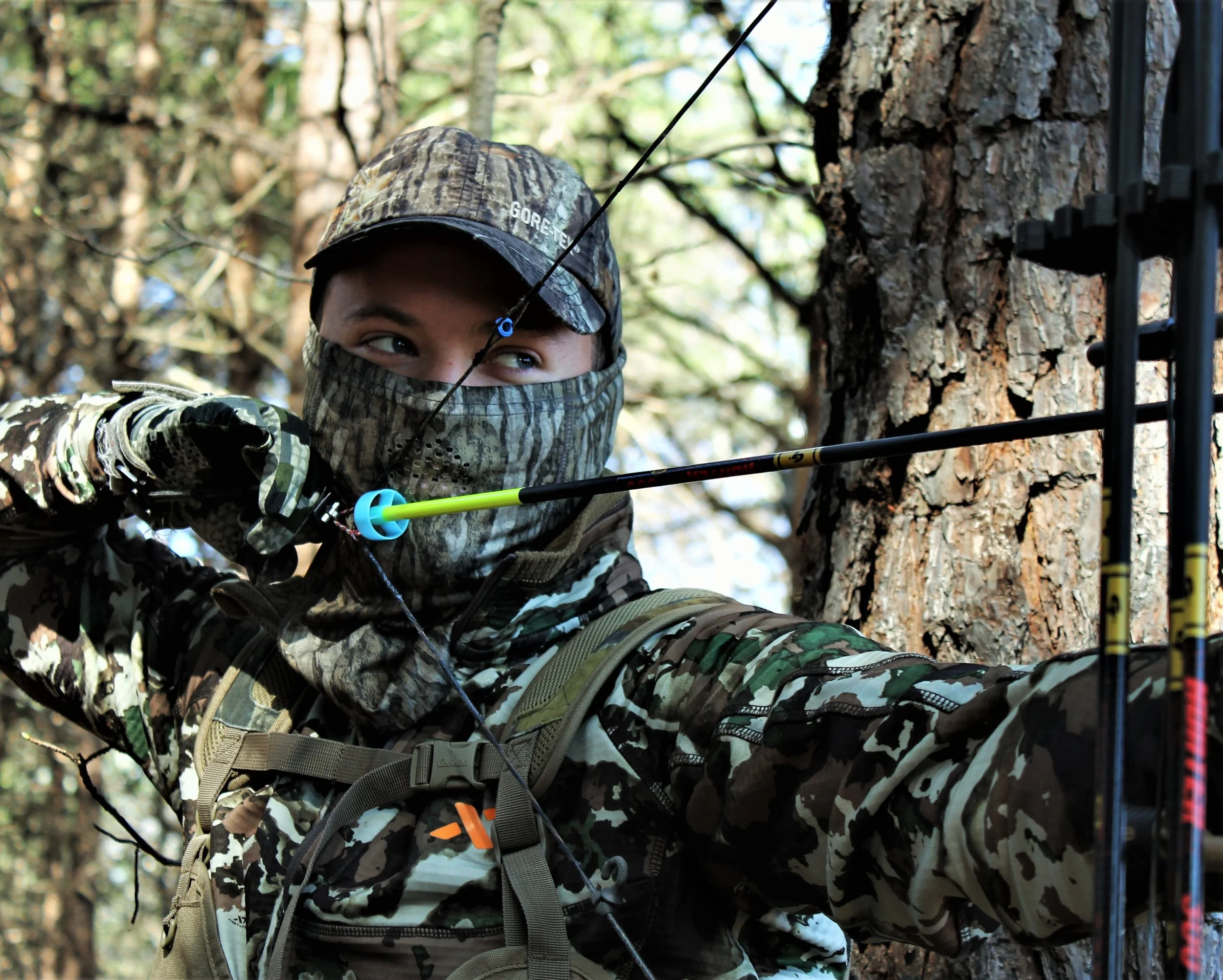 Child in camouflage hunting gear aiming a bow and arrow, partially hidden behind a tree in a forest.