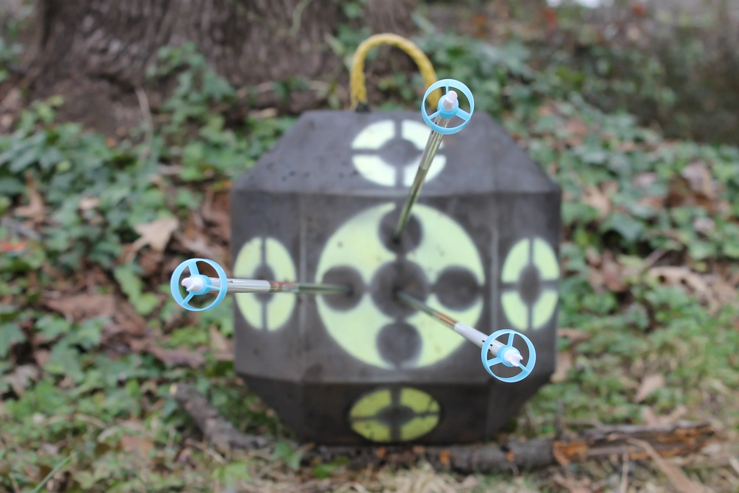 Archery target with three arrows stuck in the bullseye outdoors surrounded by grass and fallen leaves, with trees in the background.
