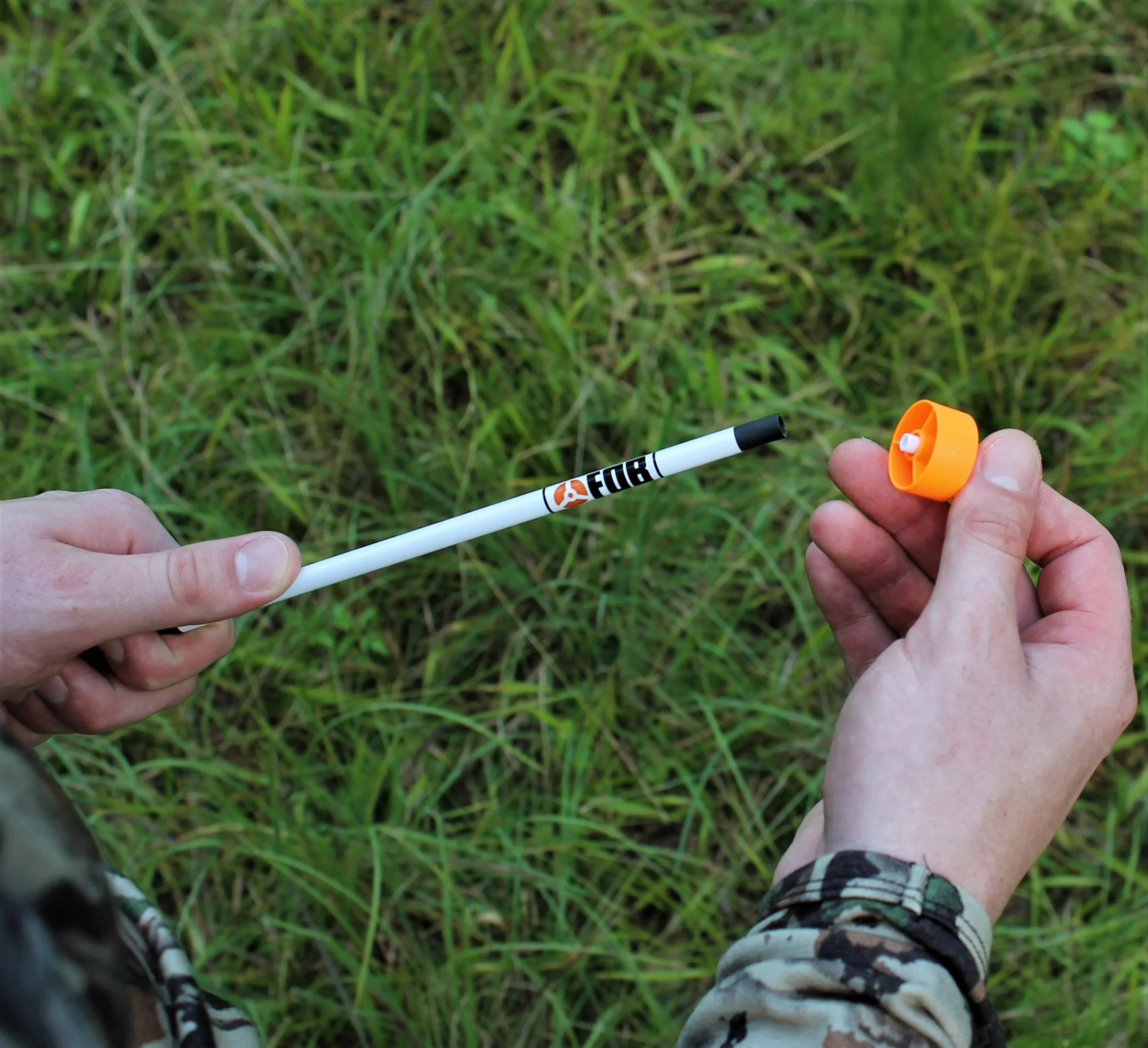 Person holding an orange and white marker with a black cap outdoors, wearing camouflage pants, in front of green grass.