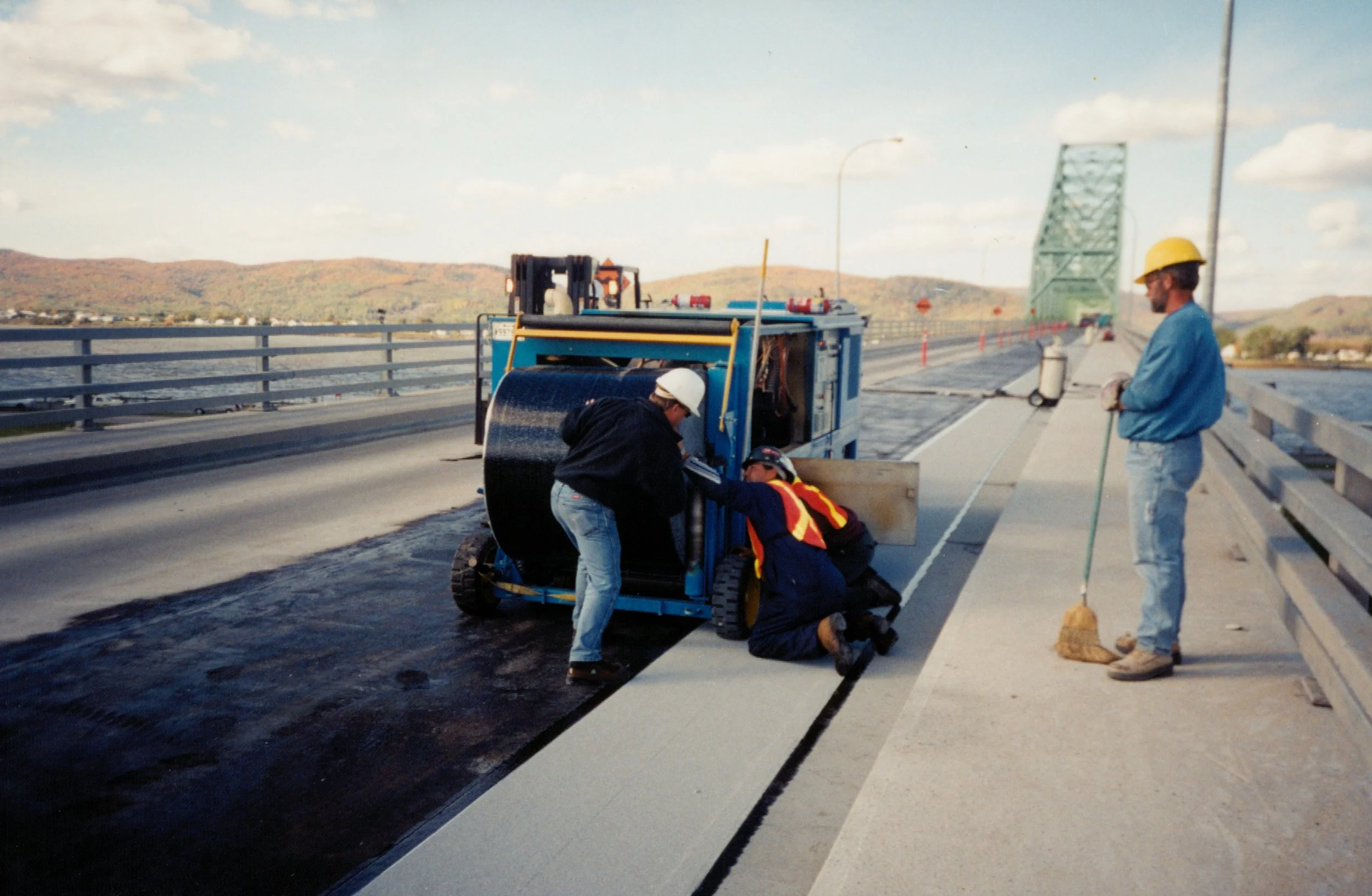 Workers laying down fresh asphalt on a bridge with a large asphalt roller, a worker operating it, and two others assisting, with a person sweeping the sidewalk nearby, and a green bridge in the background in campbellton new brunswick canada.