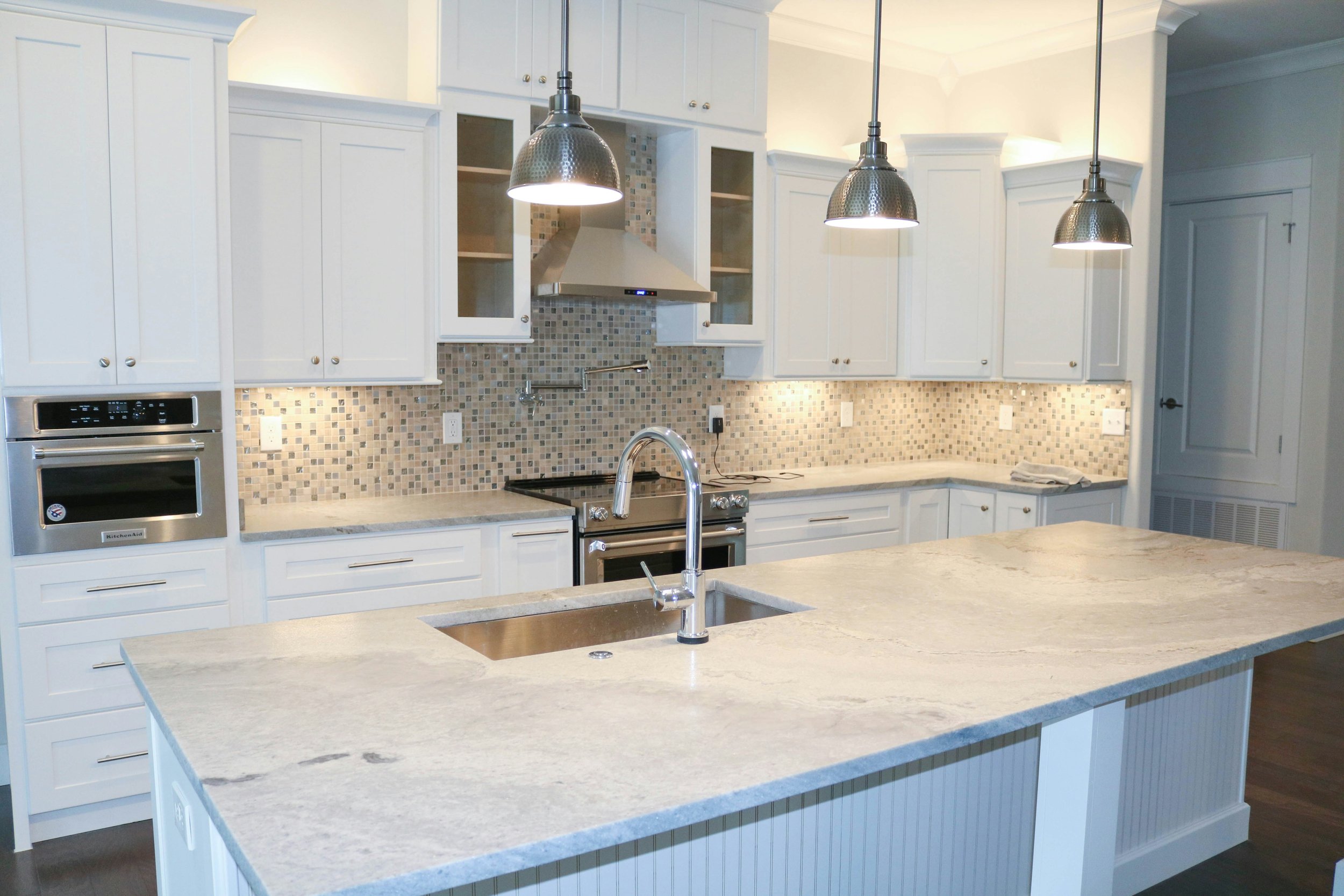A modern white kitchen with a large island, granite countertops, white cabinets, a backsplash with small mixed color tiles, pendant lights above the island, and stainless steel appliances.