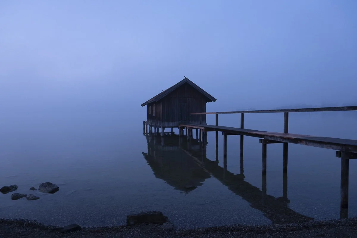 Ein Bootshaus auf Pfeilern im Wasser, mit einem Steg, bei Nebel. Silhouette einer kleinen Hütte auf dem Wasser, in ruhiger, nebliger Landschaft.