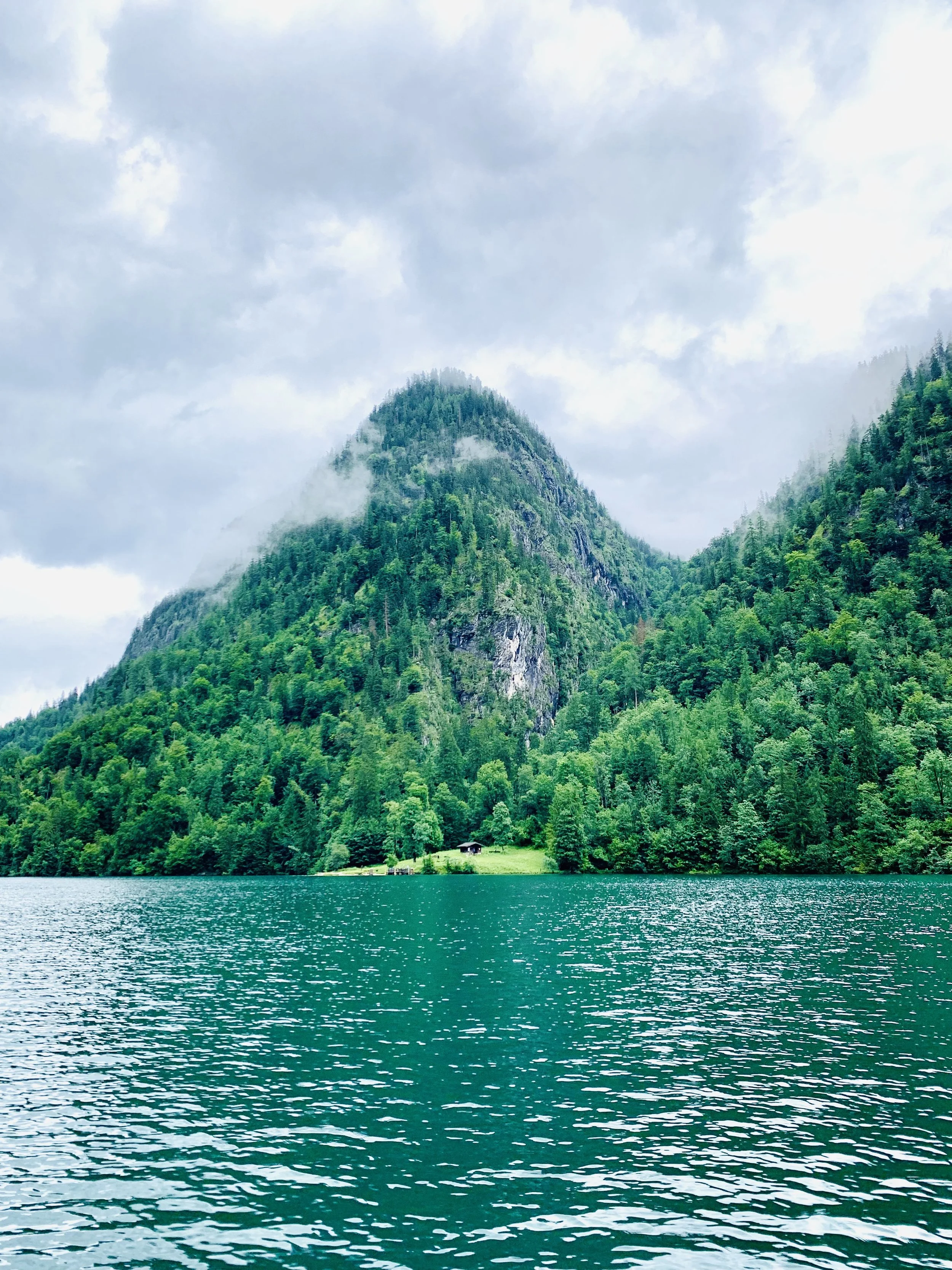 Ein See mit grün-blauem Wasser vor einem bewaldeten Berg, teils im Nebel und Wolken gehüllt, mit einem kleinen Haus am Ufer.