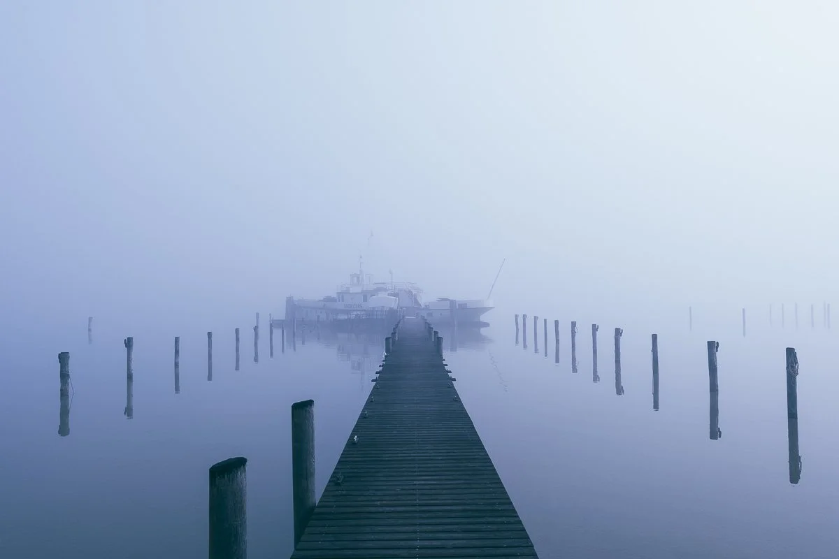 Ein hölzener Steg führt zu einem Boot auf einer ruhigen, nebligen Wasserfläche.