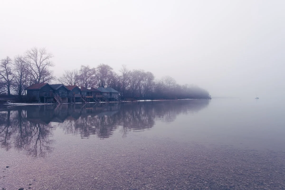 Nebel über einem ruhigen See mit Häusern am Ufer und einem Boot in der Ferne, umgeben von Bäumen.