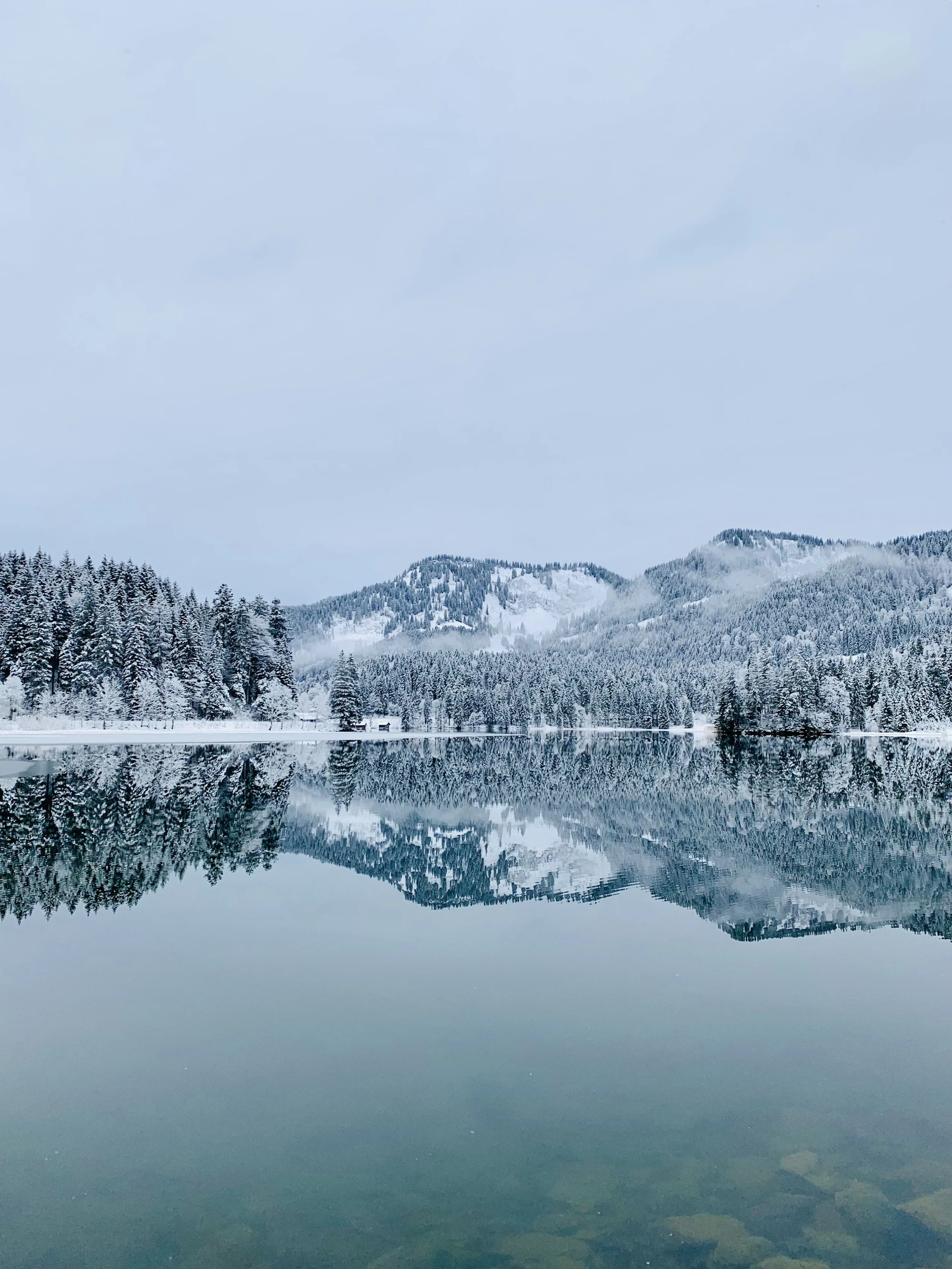 Ein verschneiter Bergsee mit gebirgiger Winterlandschaft und bewaldeten Hügeln im Hintergrund, reflektiert im ruhigen Wasser.