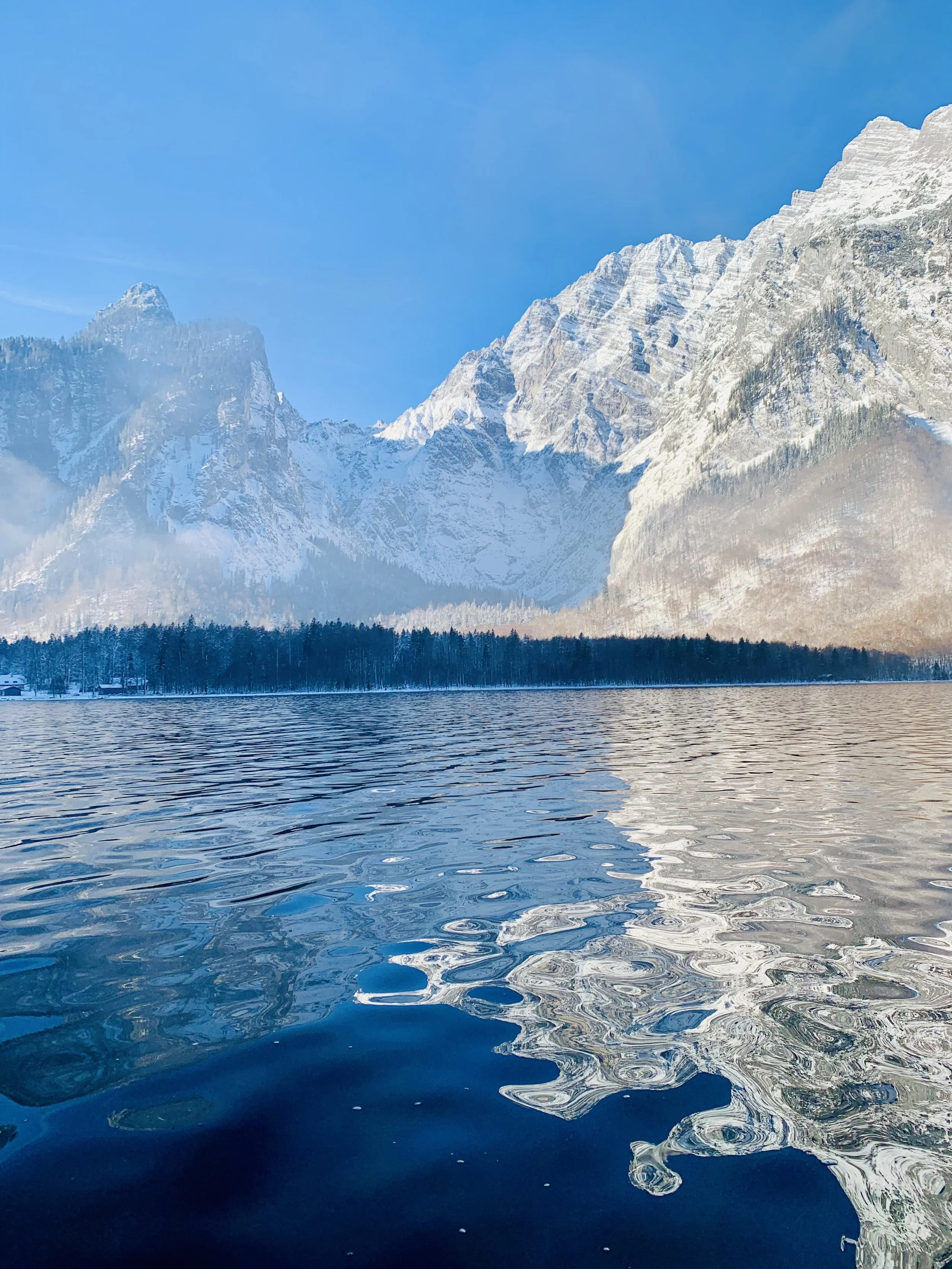 Bergsee mit schneebedeckten Bergen im Hintergrund, ein blauer Himmel und Wasser mit leichten Wellen im Vordergrund.