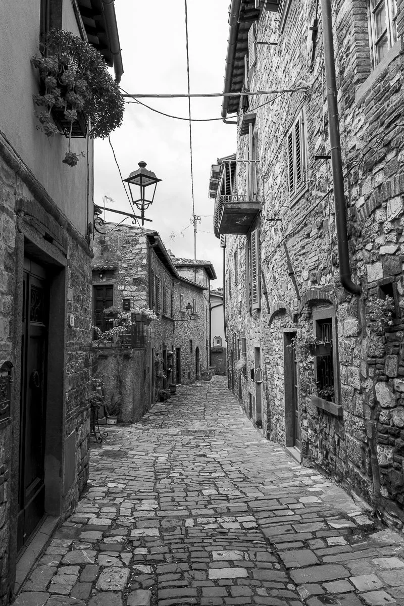 Eine enge, kopfsteingepflasterte Gasse mit alten, steinernen Gebäuden auf beiden Seiten, mehrere Fenster mit Blumen und ein Laternenlampen hängen über der Straße.