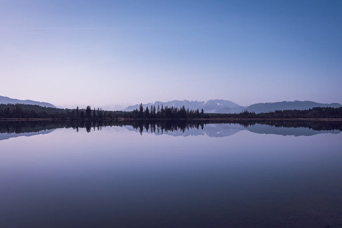Eine ruhige Seenlandschaft mit Bergen im Hintergrund und Wald entlang des Ufers, alles spiegelt sich im Wasser wider.
