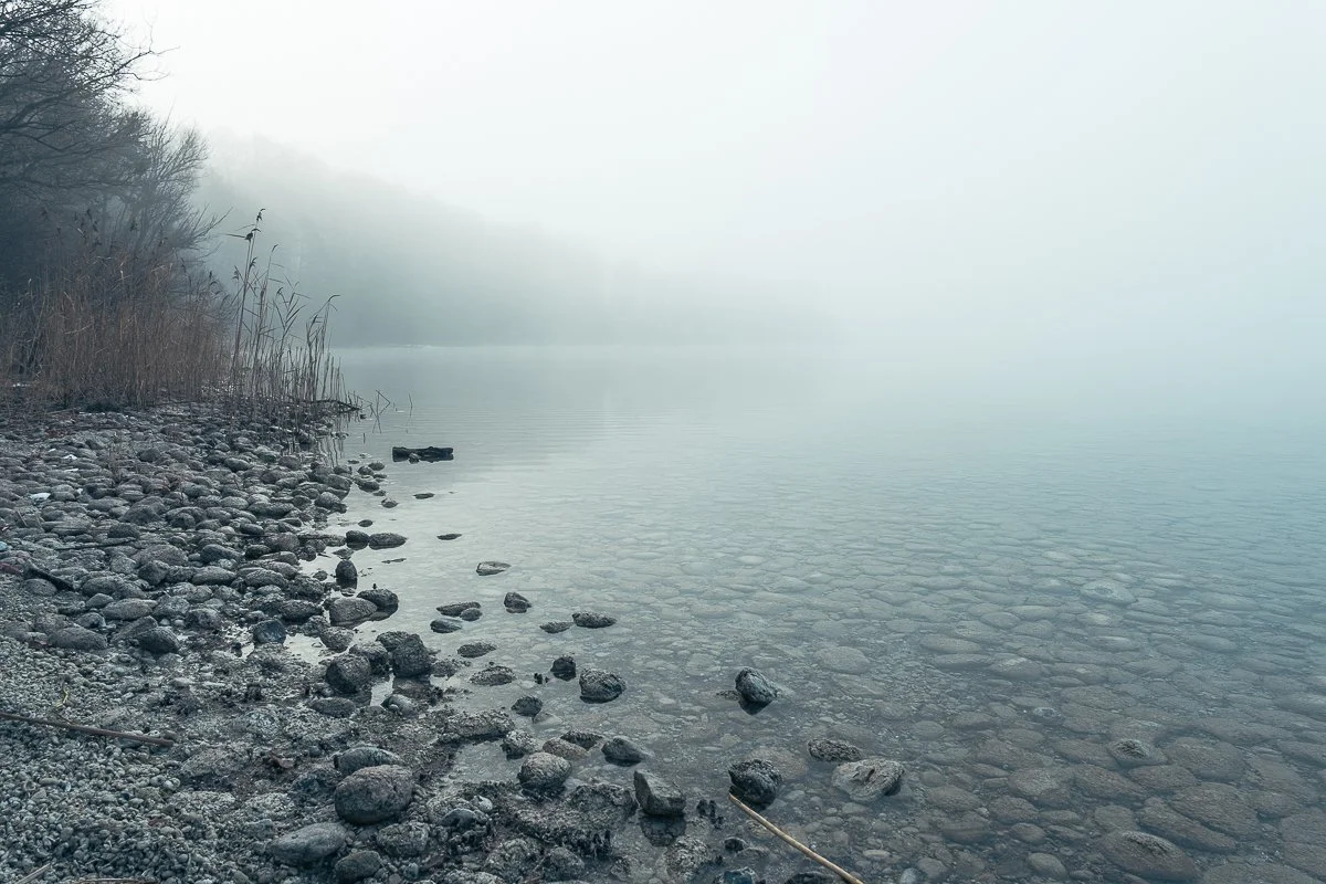 Nebelverhüllter See mit felsigem Ufer und Khaki-Grauen Vegetation am Rand.