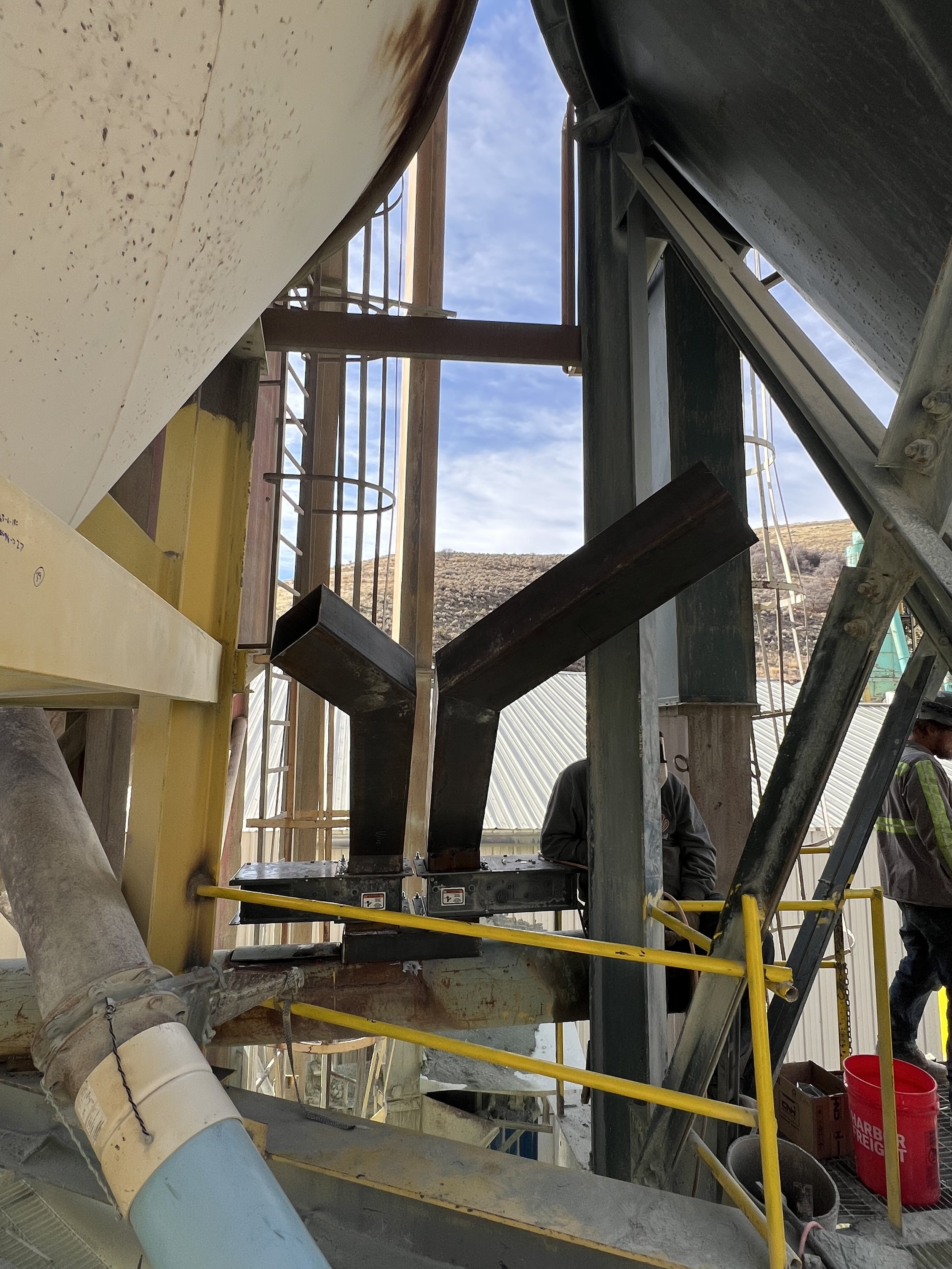 Construction site with steel framing and workers, outdoors with a mountain and partly cloudy sky in the background.
