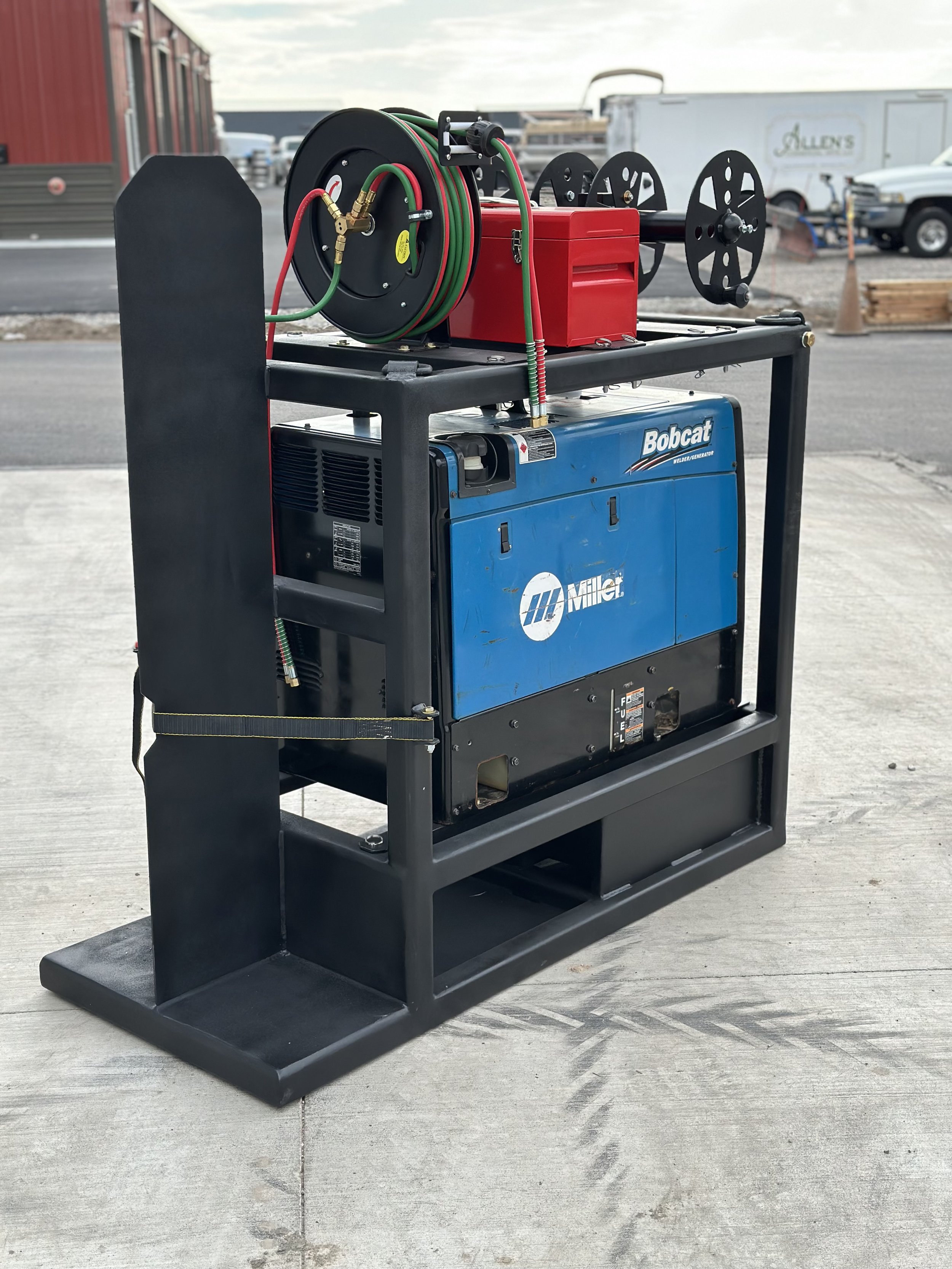 A mobile welding machine with a blue Miller Bobcat generator, a reel of welding hose, and some other accessories mounted on a black metal frame, outdoors on concrete.