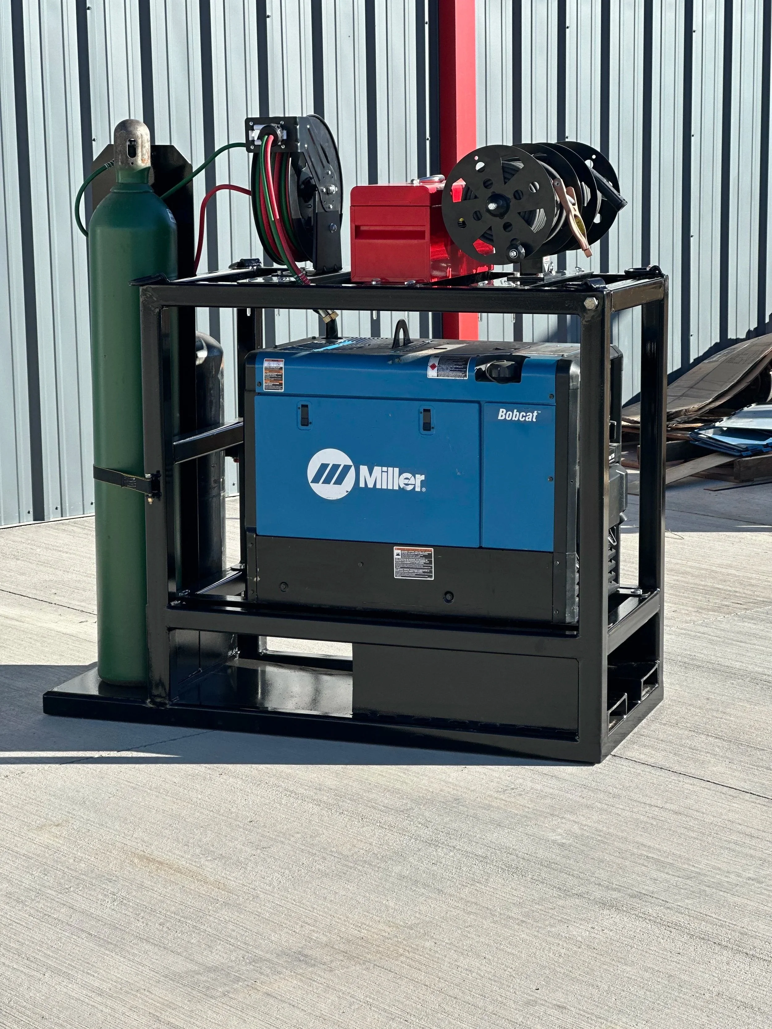 Portable generator setup with a blue Miller generator, a green oxygen tank, and red compressor on a black metal frame outside a building.