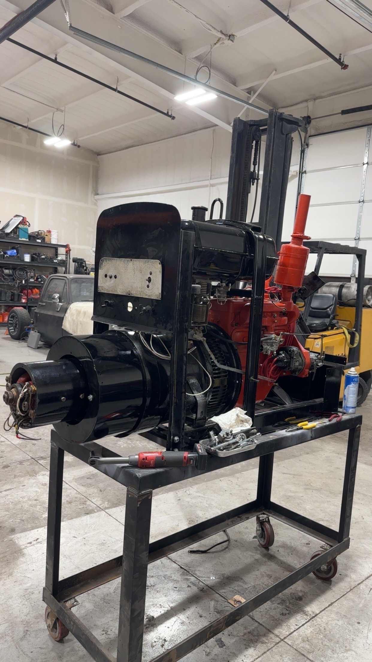 A large mechanical device or engine on a rolling cart inside a workshop or garage, with various tools and equipment in the background.