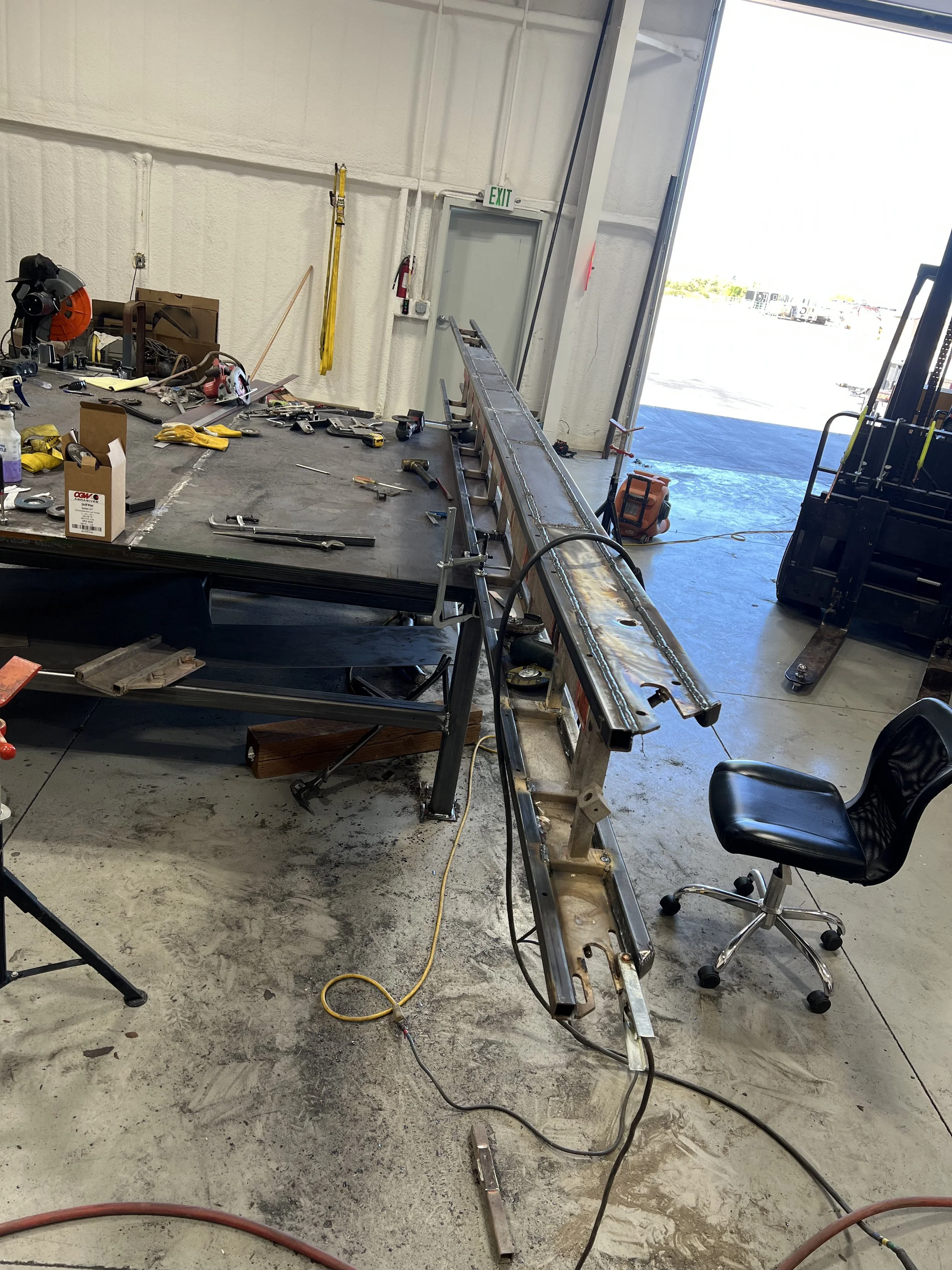 Inside a workshop with a long metal frame, tools, a black chair, and an open garage door showing a parking lot outside.