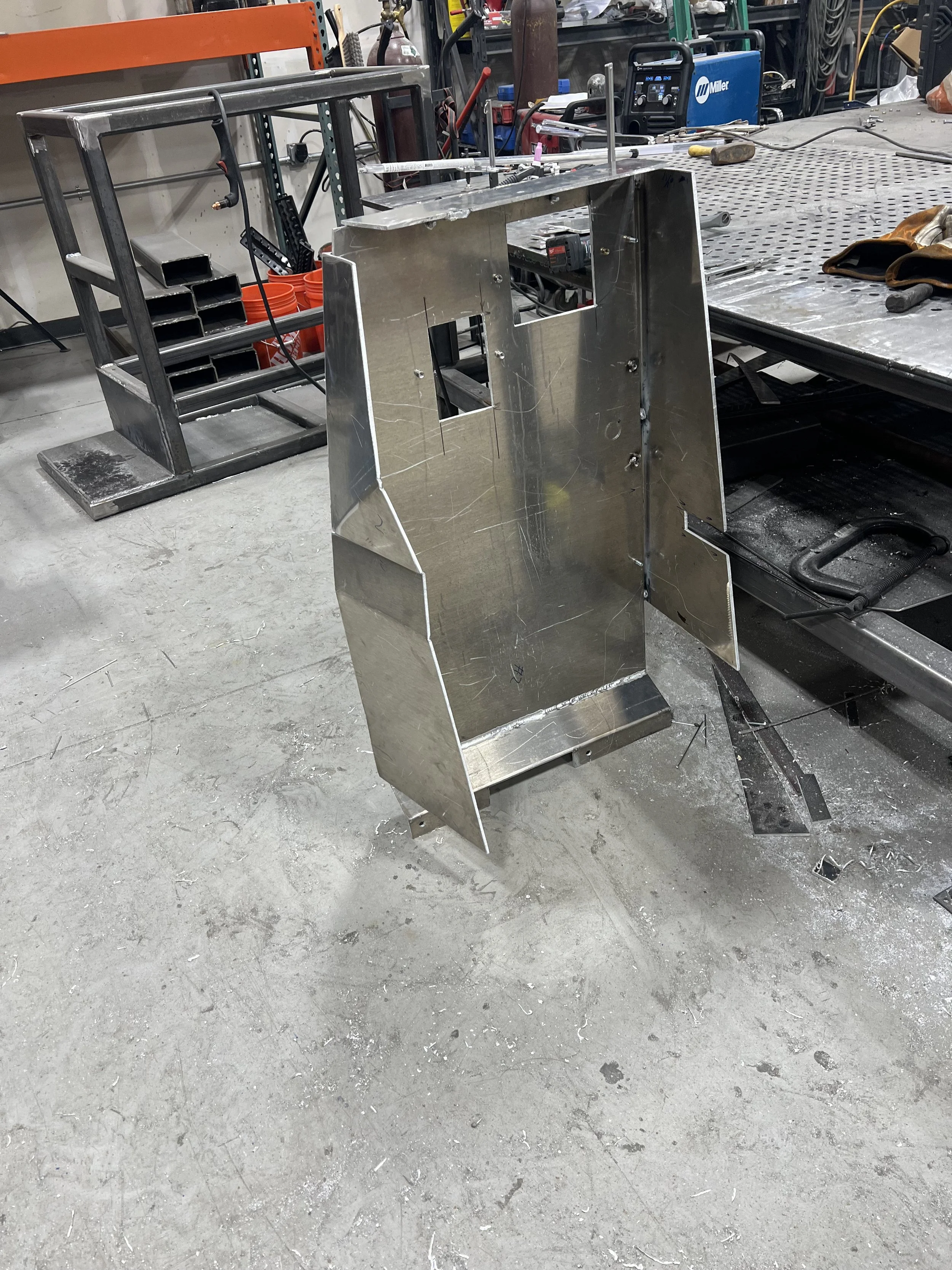 Metal fabrication workshop with a large, unfinished metal structure in the foreground, a workbench with tools, and welding equipment in the background
