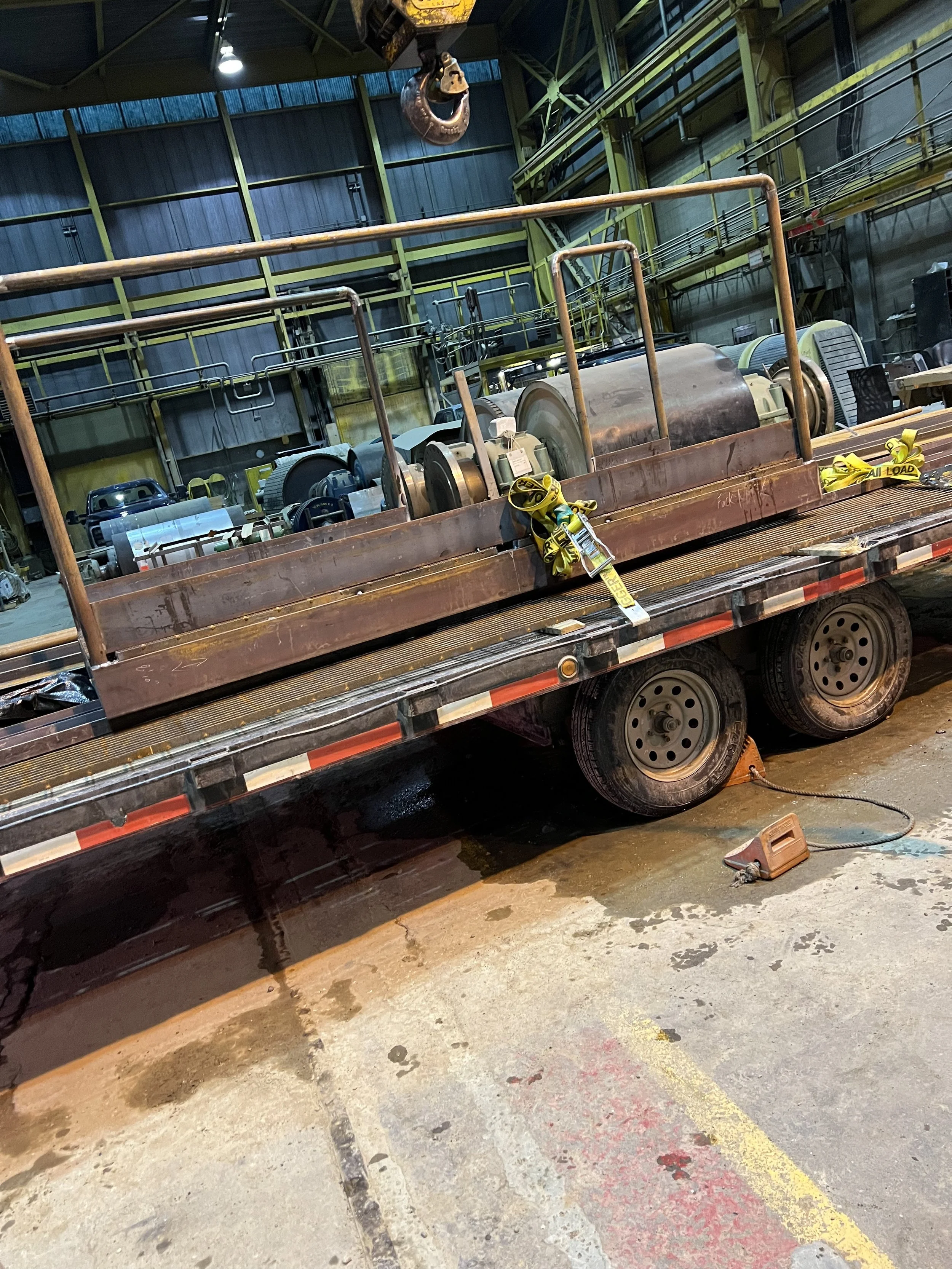 A flatbed truck inside an industrial warehouse, carrying a large industrial machine with metal frames and wheels, yellow straps securing the machine, and a floor with oil stains and a power cable.