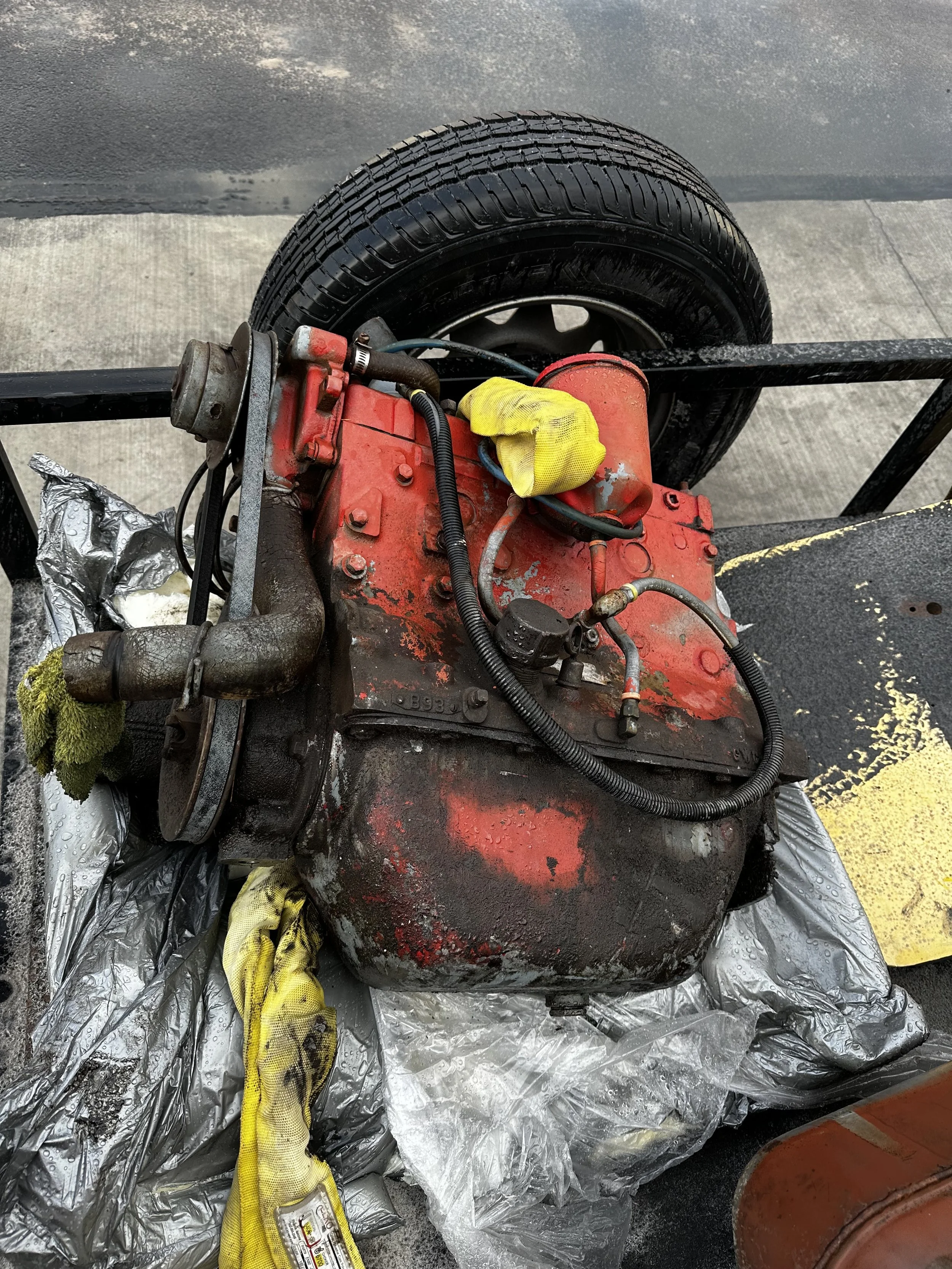 An old, red engine with dirt and rust, resting on a silver plastic sheet, with a tire and pavement in the background.