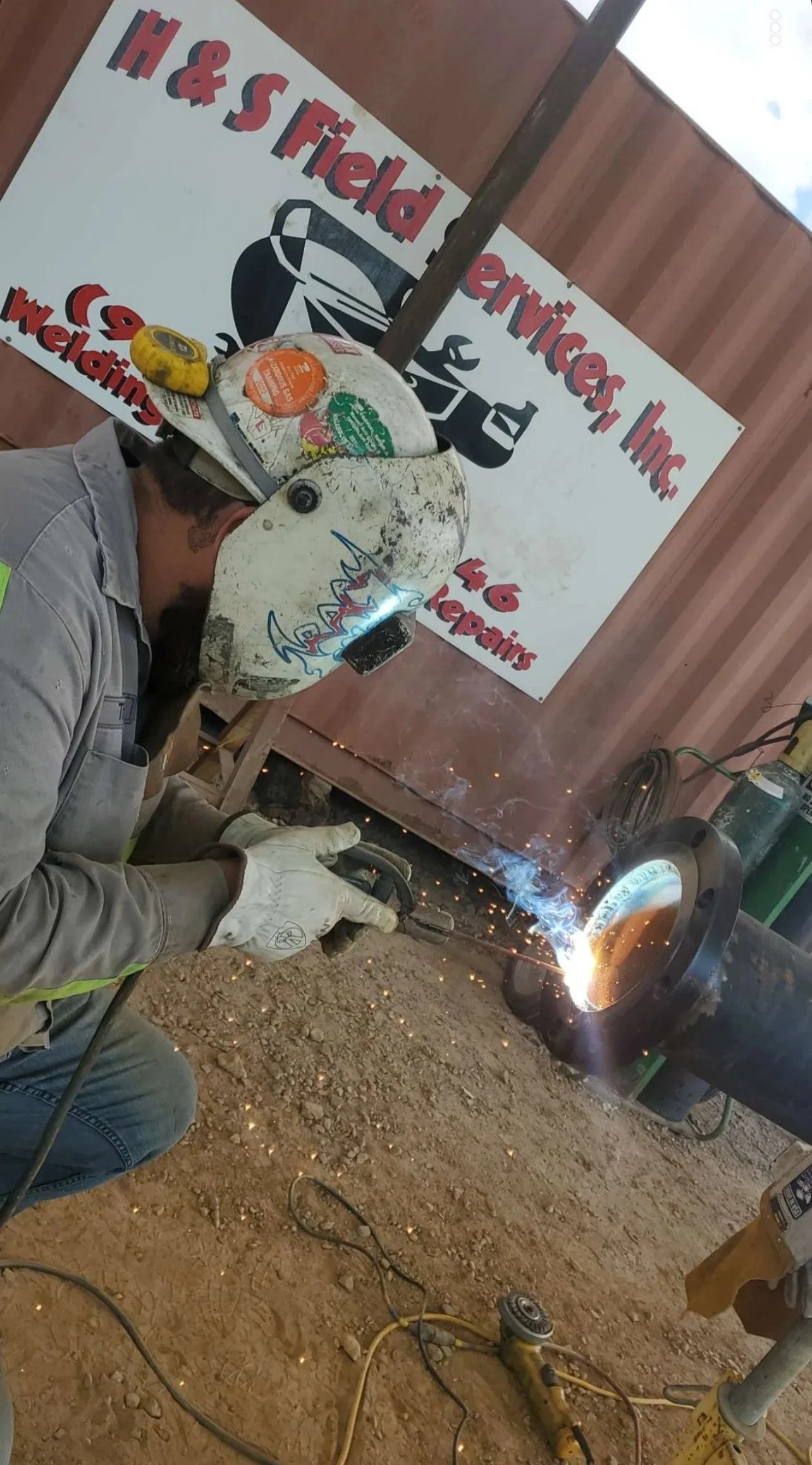 A worker welding a pipe at a construction site, with sparks flying. The worker is wearing a safety helmet and gloves, and there is a sign in the background for H & S Field Services, Inc.
