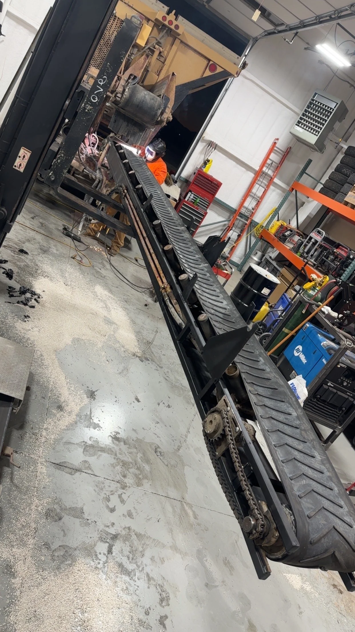 A person welding inside a workshop with a large conveyor belt or chain system extending across the image, surrounded by tools, equipment, and industrial supplies.