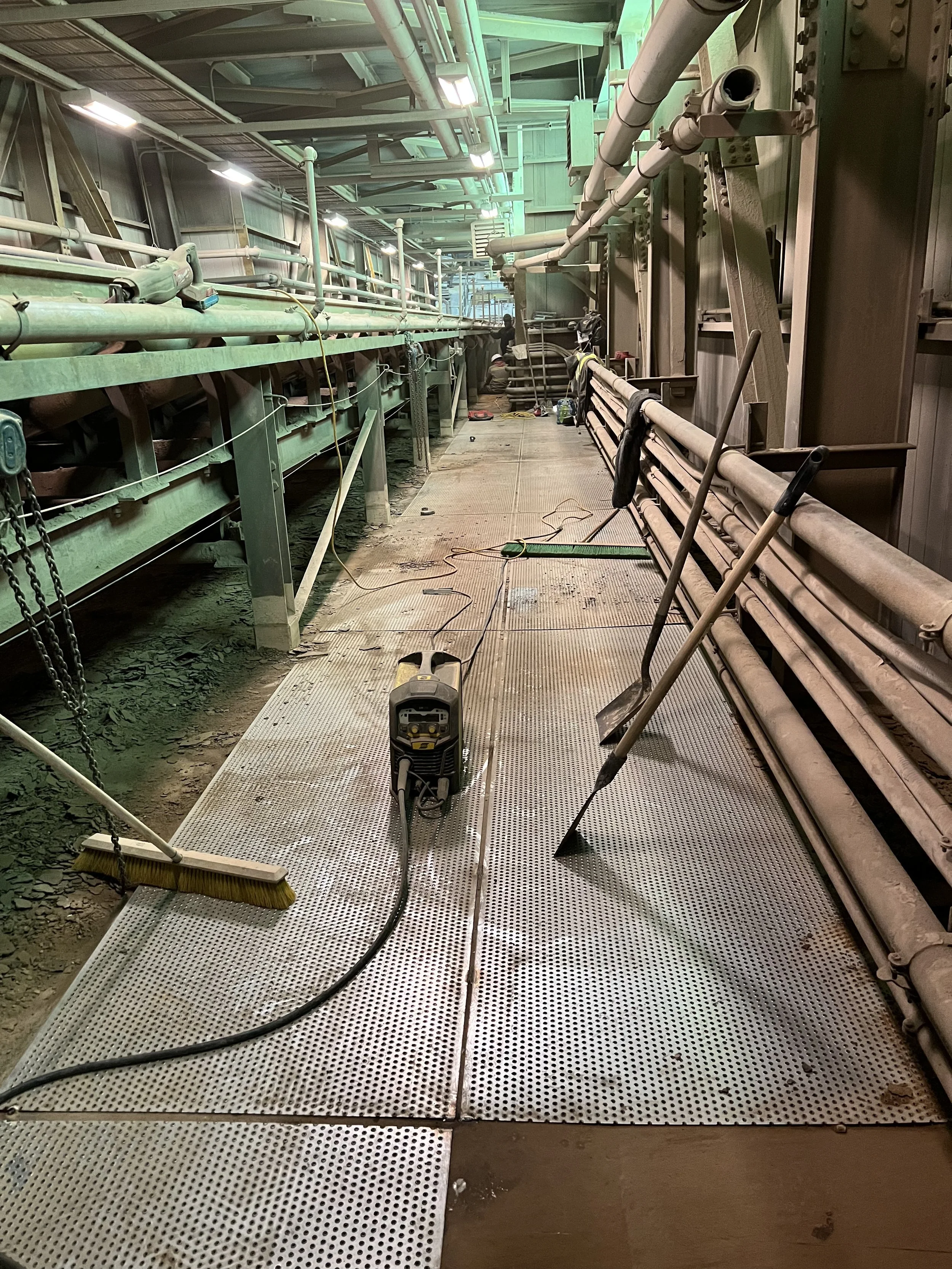 Industrial workshop with metal flooring, tools leaning against the wall, and a small electronic device with wires on the ground. The area is cluttered with pipes, hoses, and equipment along the walls.