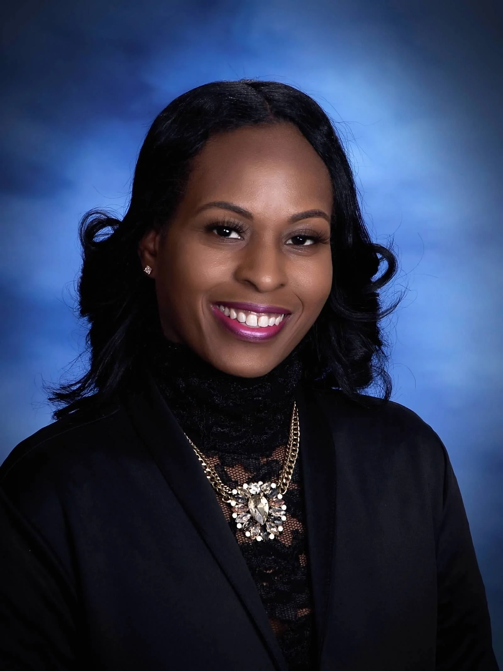 portrait of a smiling African American woman with black wavy hair, wearing a black top, a statement necklace with a large decorative gemstone, and earrings, against a blue background.