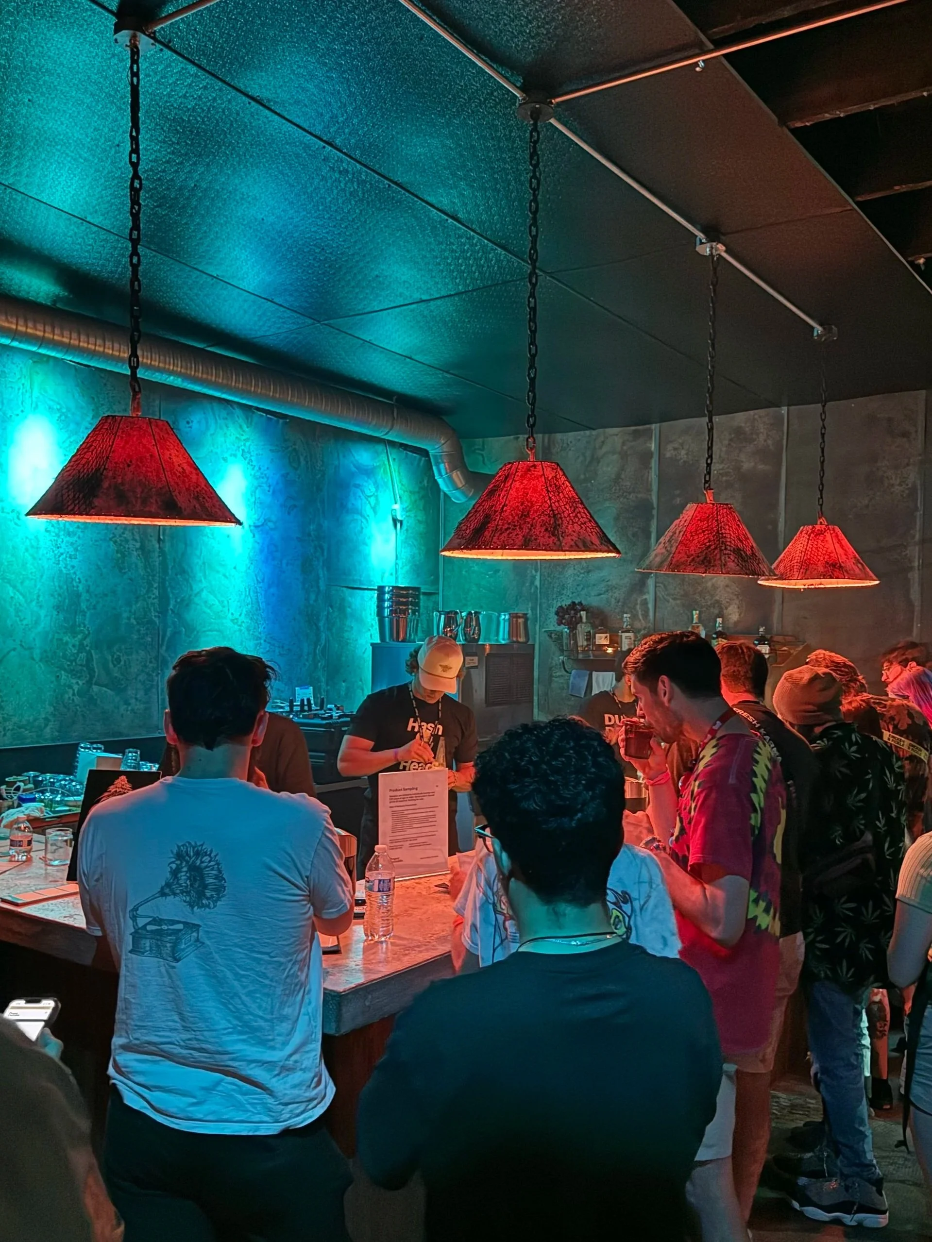 People standing around a bar counter with neon lighting and red hanging pendant lamps in a dimly lit bar or club.