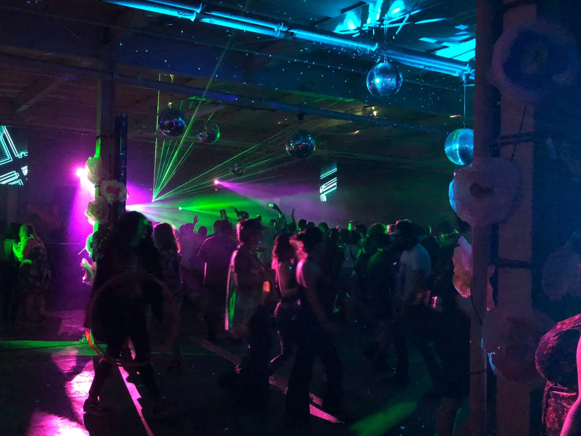 People dancing at a nightclub with colorful lights, including green lasers, purple and blue spotlights, and disco balls hanging from the ceiling.