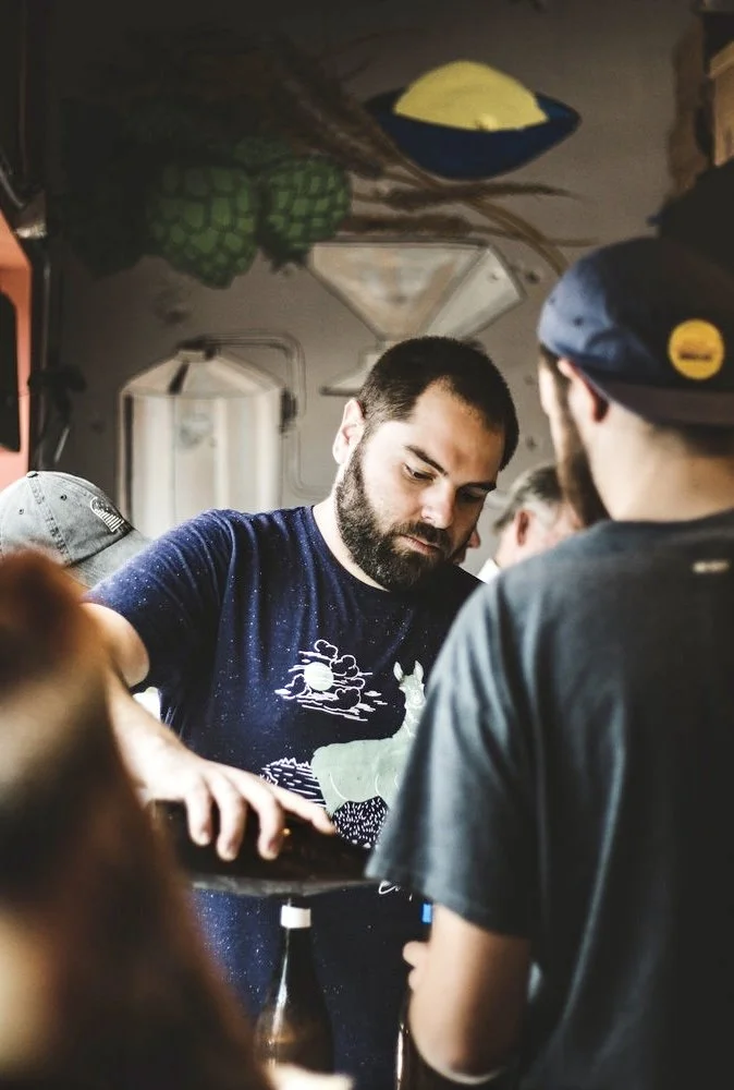 A man with a beard wearing a navy t-shirt with a graphic of a sunset over water, serving drinks at a social gathering. Several other people are around him in a casual setting, with a background of a painted mural featuring green grapes and abstract shapes.