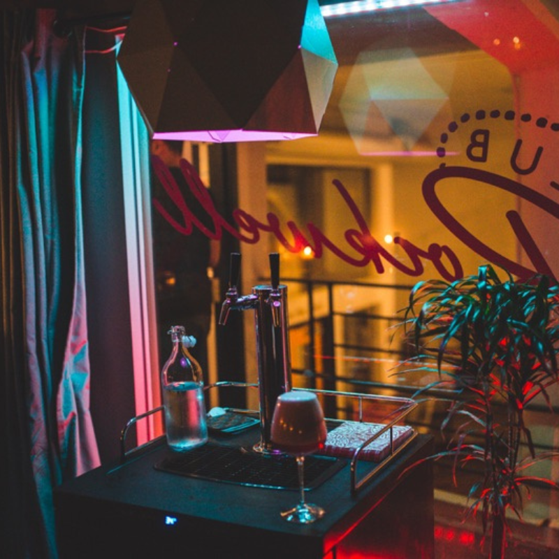A bar area with a colorful light fixture, a glass of wine, a water bottle, a book, and a plant, seen through a window with red cursive writing.