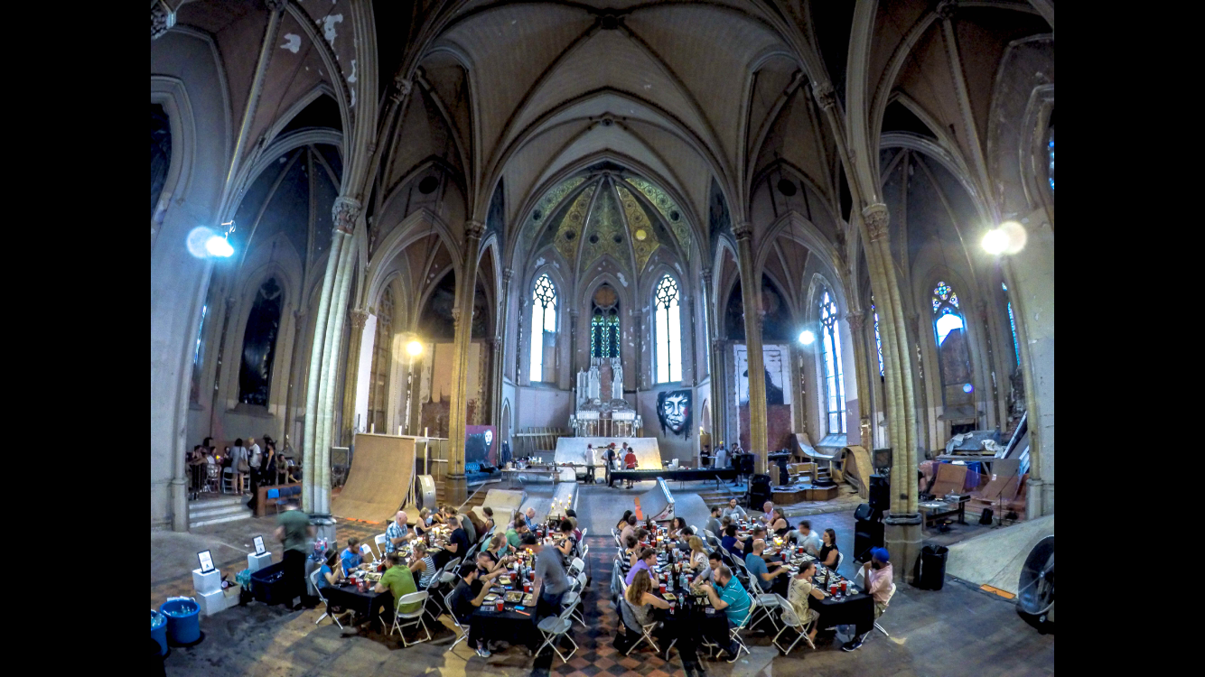 People sitting at tables inside a large gothic-style cathedral with stained glass windows, high vaulted ceilings, and artwork, participating in an event or gathering.