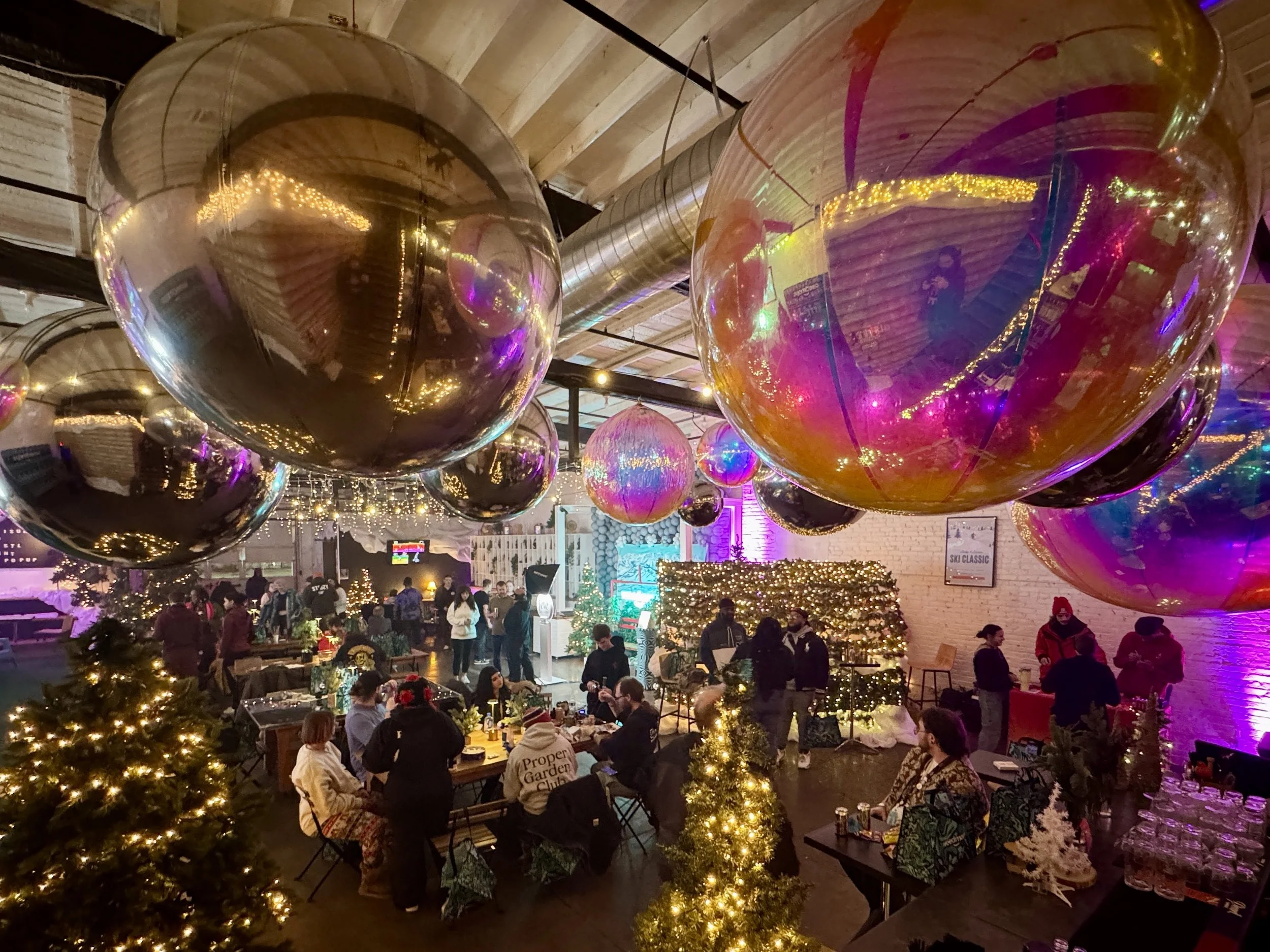 Decorated indoor space with large reflective balloons hanging from the ceiling, string lights, small Christmas trees, and people socializing and dining.