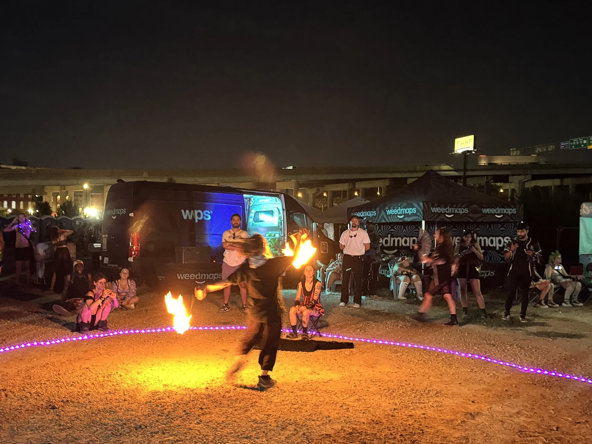 A nighttime outdoor event with a fire performer in the foreground and a crowd watching, under a dark sky with city lights and bridges in the background. The event is set up with weedmaps tents and a vehicle with branding.