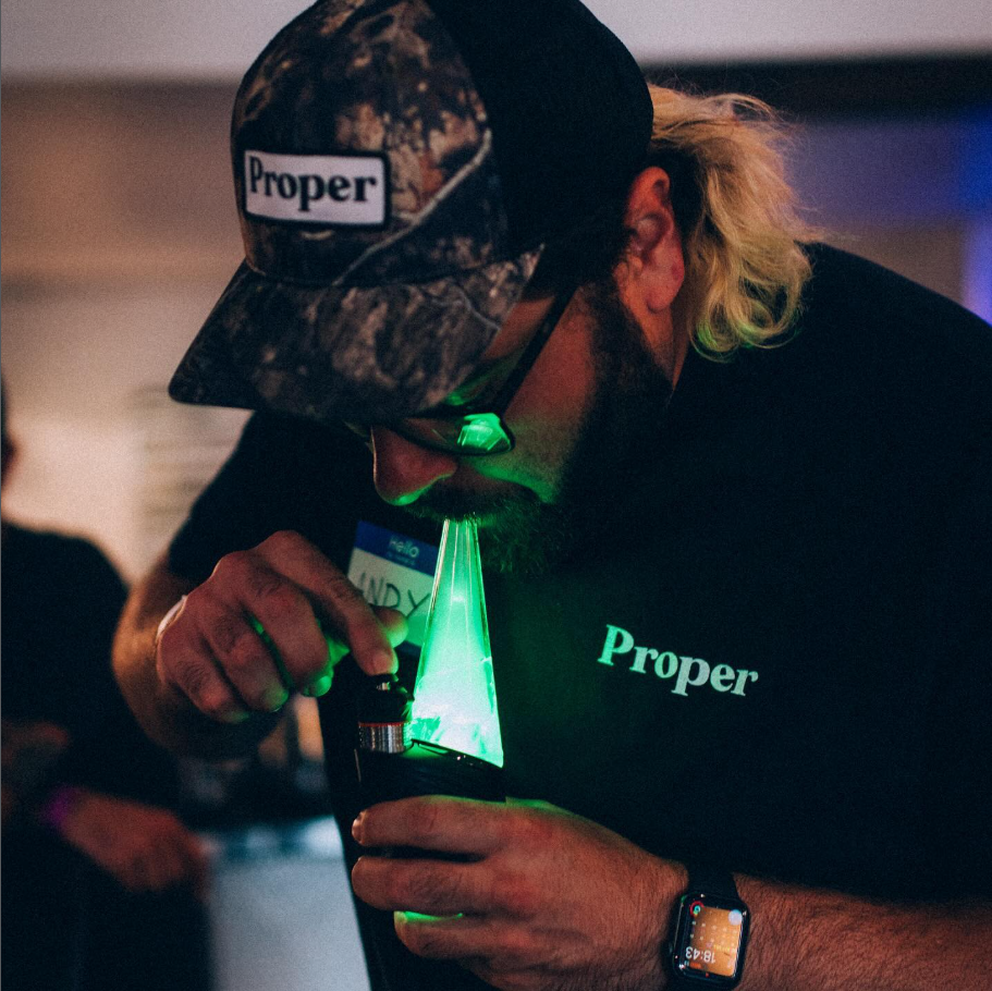 Person with long blond hair, cap labeled "Proper", and smartwatch, examining a green laser beam through a microscope at a conference or event.