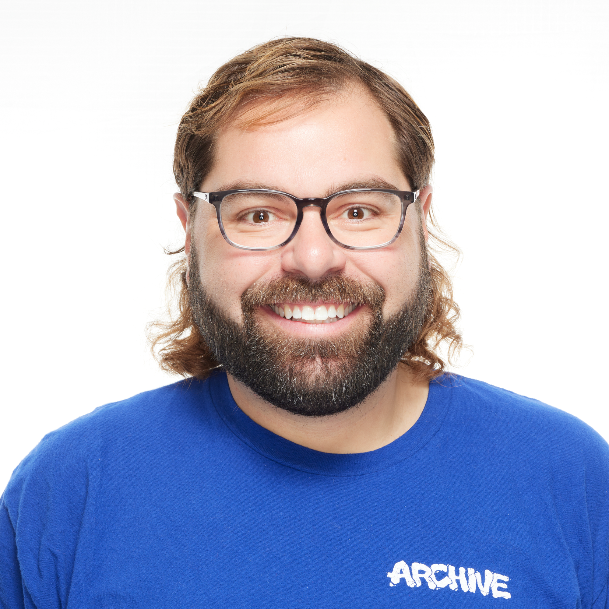 A smiling man with glasses, a beard, and long hair wearing a blue shirt that says 'ARCHIVE'.