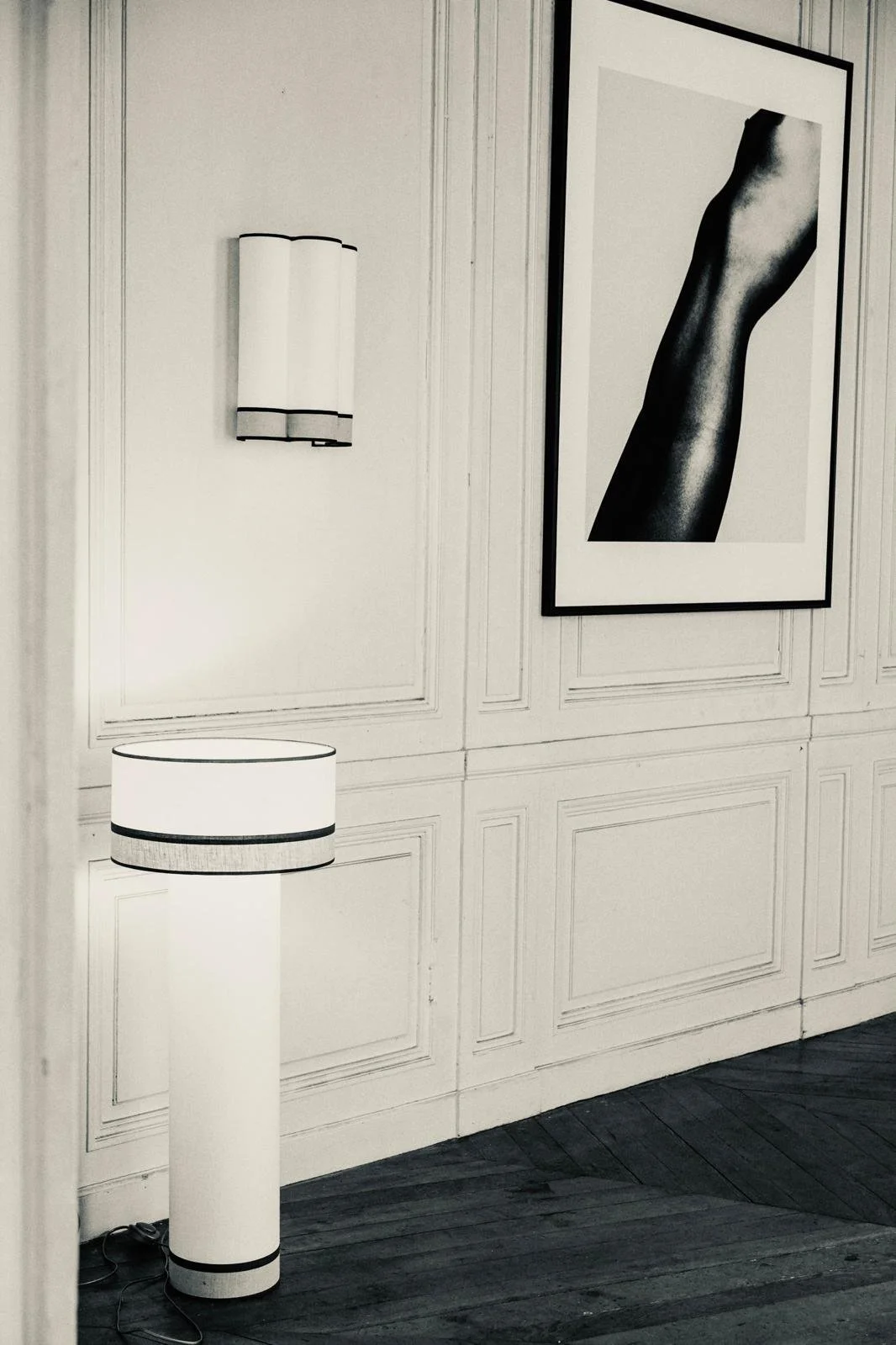 Photo en noir et blanc d'une pièce avec un tableau représentant une jambe nue et deux lampes modernes, une murale et une à terre, dans une décor classique avec panneaux en bois sur le mur.