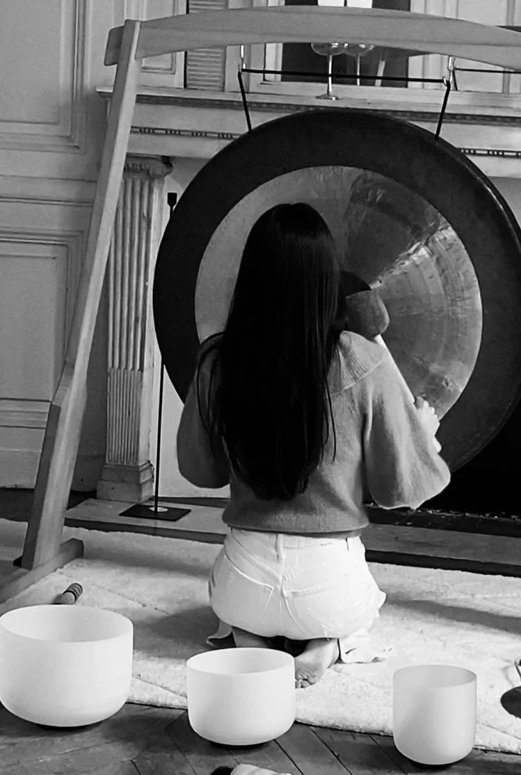 Une personne avec de longs cheveux noirs, vue de dos, assise à genoux devant un gong doré dans une pièce élégante. Trois bols chantants en avant et un trapèze au-dessus du gong.