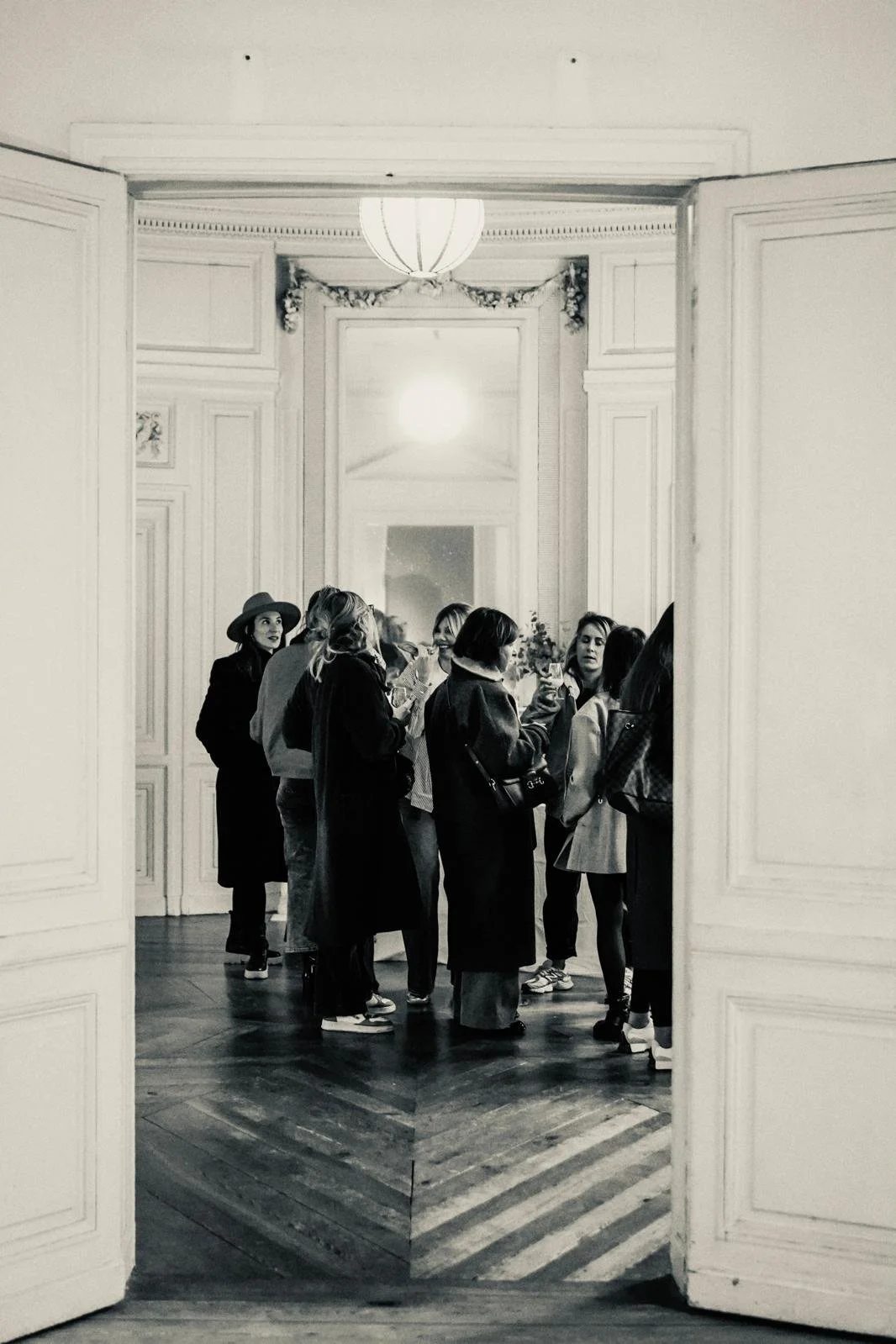 Groupe de personnes debout dans une pièce élégante au fond d'une pièce, vue à travers une porte ouverte, en noir et blanc.