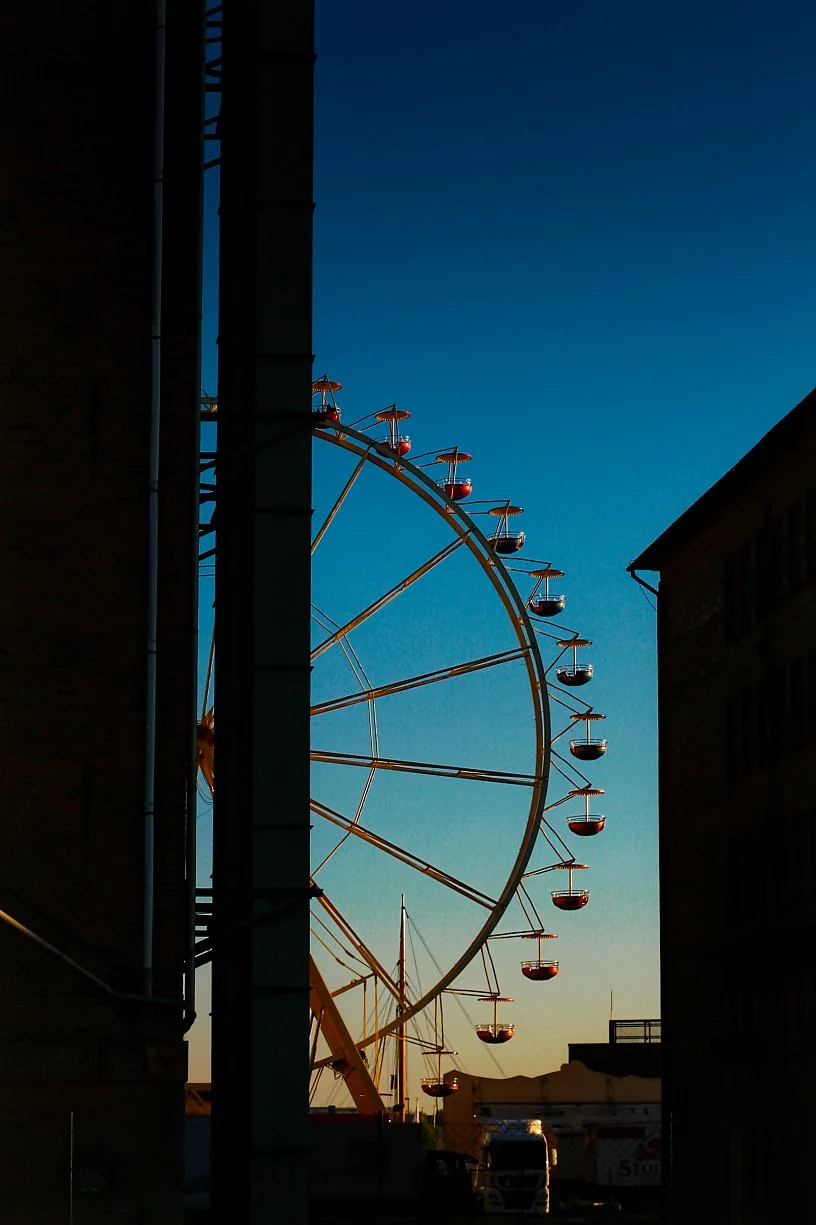 Silhouette of a Ferris wheel against a dusky sky, partially obscured by dark buildings.