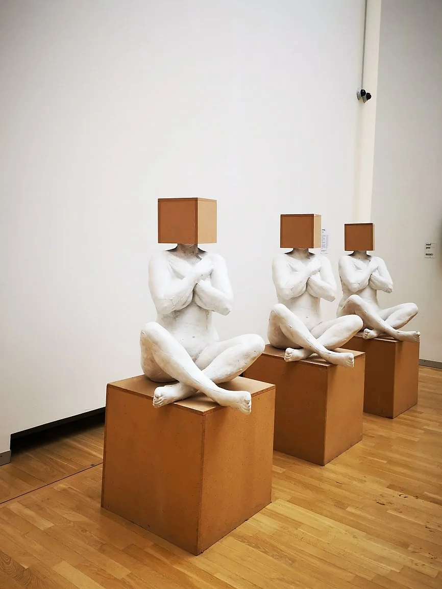 Tre kunstneriske skulpturer av nakne mennesker med bokser som hodet, plassert på tre tre-skrin i et galleri med trebenet gulv.