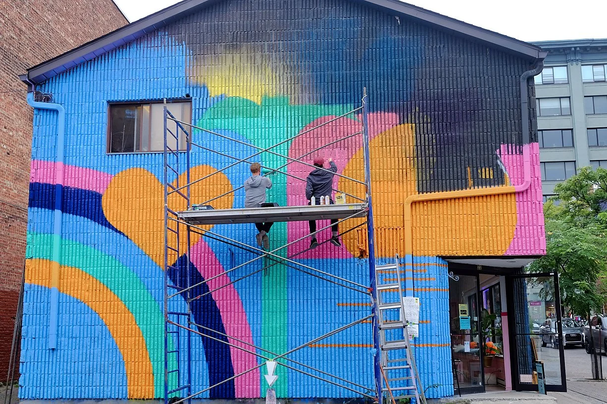 Two people on scaffolding painting a colorful mural on the side of a building with geometric shapes and bright colors.