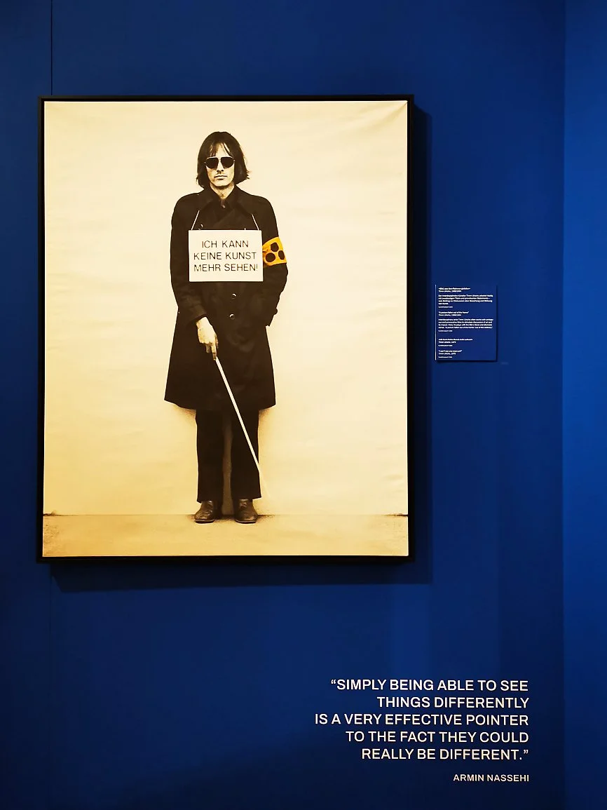 Fotografi av en person med mørkt hår, solbriller og svart klær som holder en signatur som sier "Ich kann keine Kunst mehr sehen!". Personen står på en beige bakgrunn. Bildet er utstilt i et galleri med blå vegg og et plakett ved siden av.