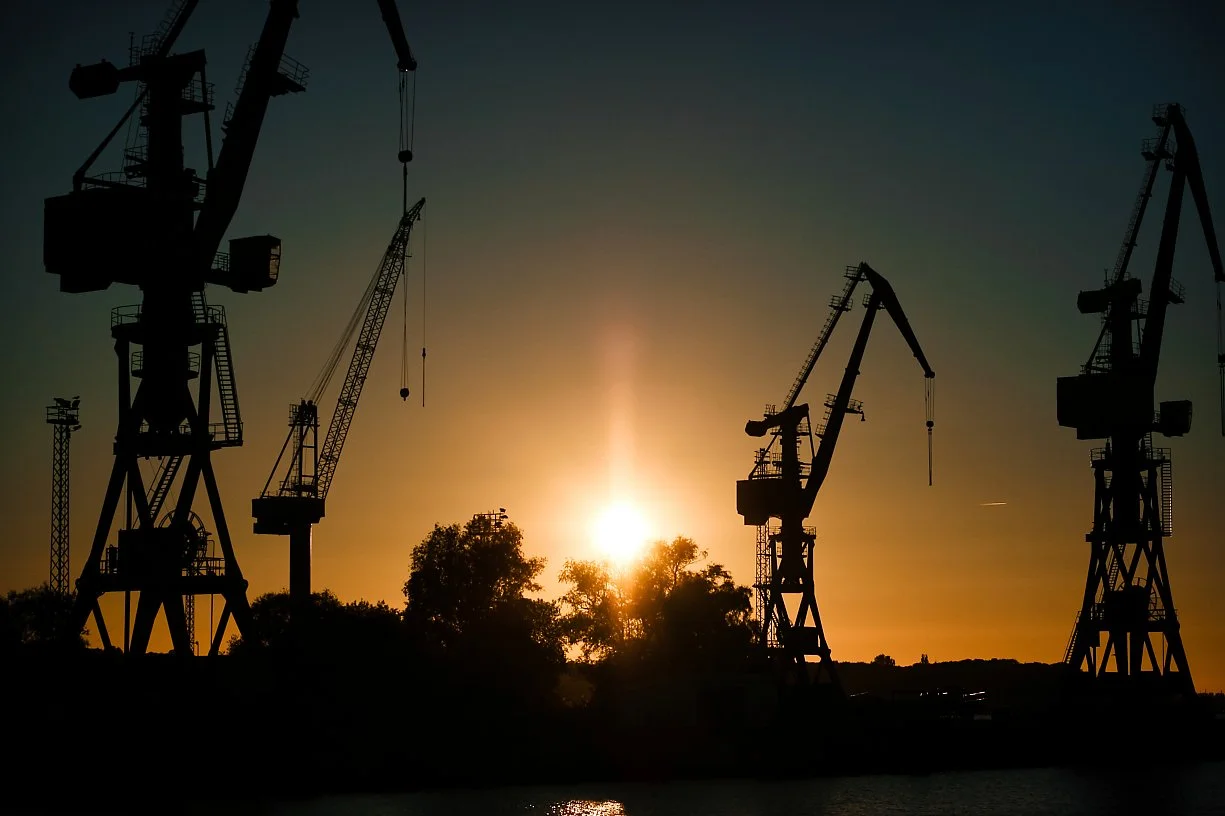 Silhouettes of large industrial cranes at sunset or sunrise with the sun near the horizon and trees in the background.