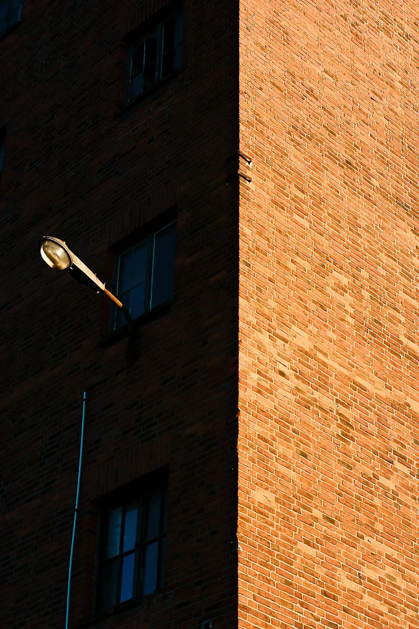 Close-up of a brick building showing two windows, a streetlamp, and a shadow cast by the building.