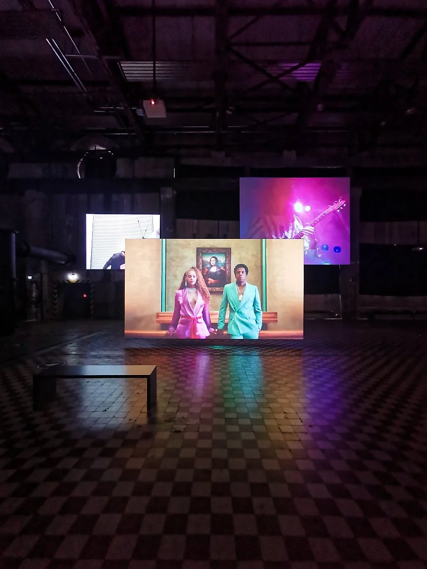 Et mørkt rom med flere skjermer som viser forskjellige bilder. En av skjermene i midten viser to personer i fargerike dresser foran et maleri. En annen skjerm til høyre viser en person med elektrisk gitar under lysende scenelys.