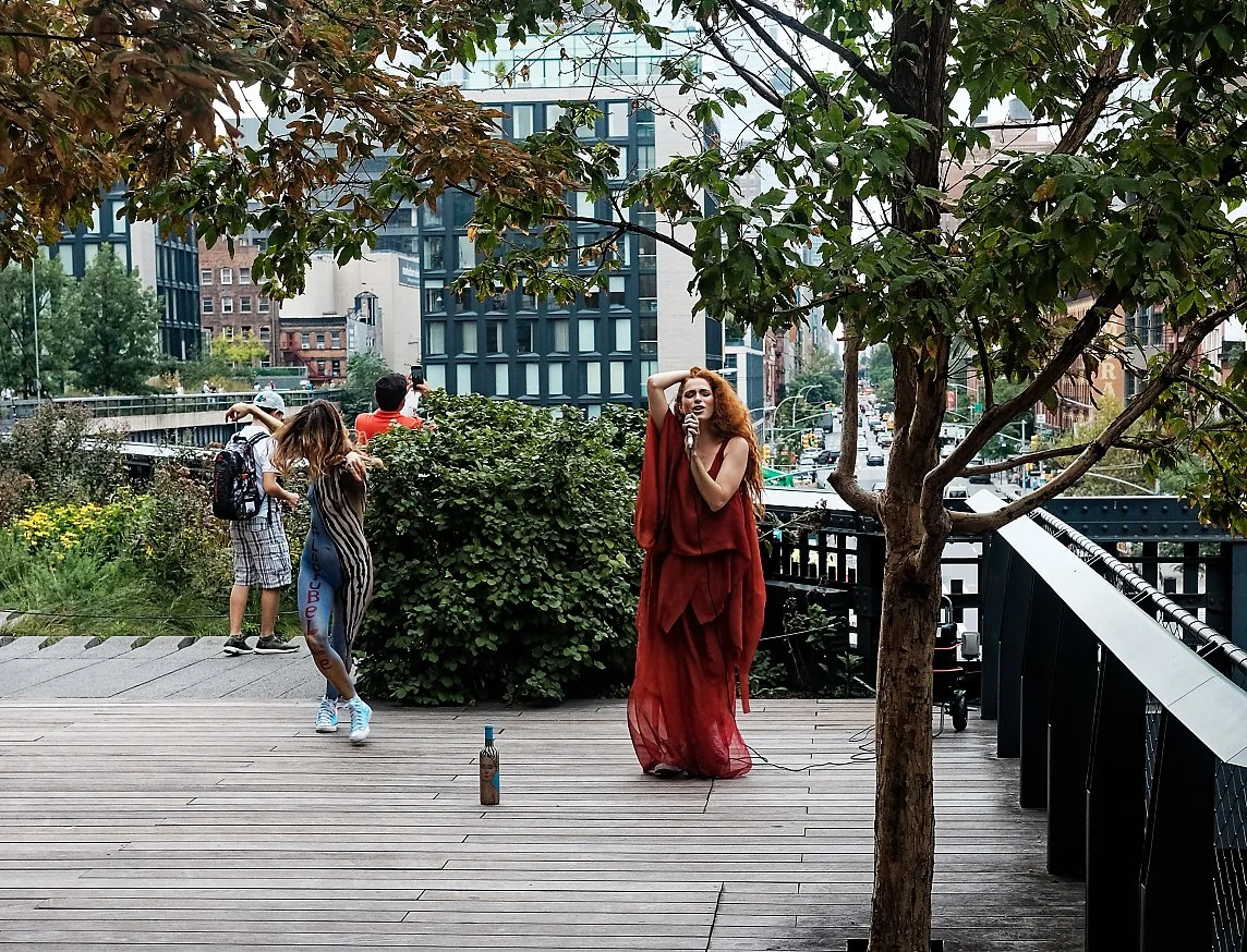 A woman singing into a microphone on an outdoor wooden stage, with a small group of children nearby and urban buildings in the background.