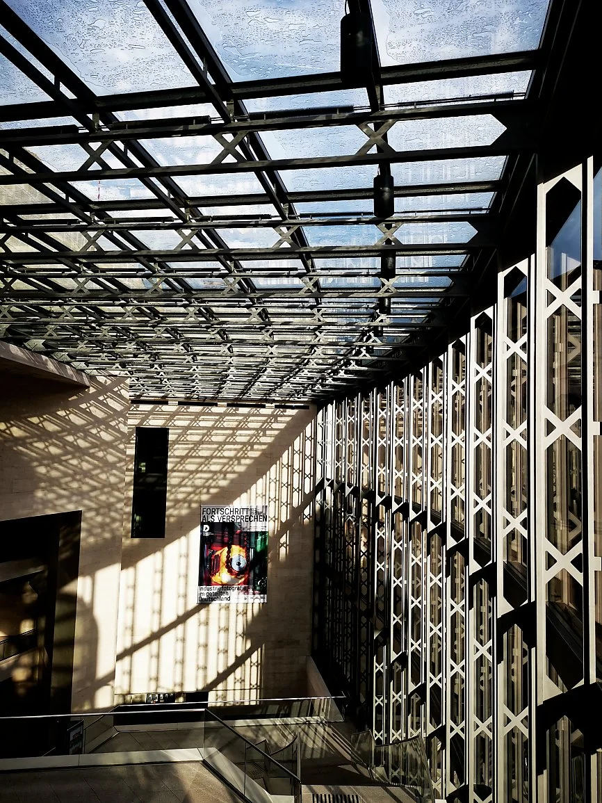 Innendørs område med glasstak og store vindusflater med utsikt mot himmelen, skygger av takrammer på veggen, og en plakat med tekst.