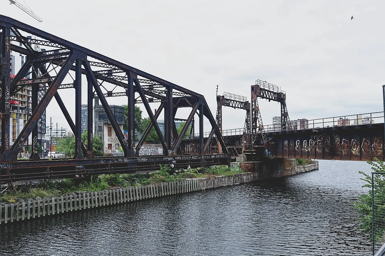 An old, rusted drawbridge over a body of water in an urban area with graffiti on the bridge structure and buildings in the background.