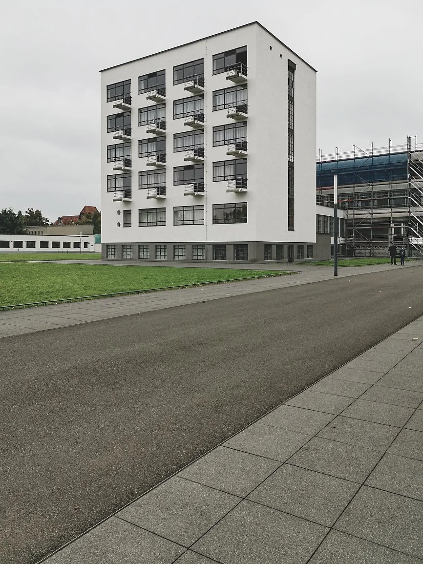 A modern multi-story apartment building with several balconies and large windows. The building is white with dark frames. There is a paved walkway and green grass in the foreground, with an overcast sky overhead.
